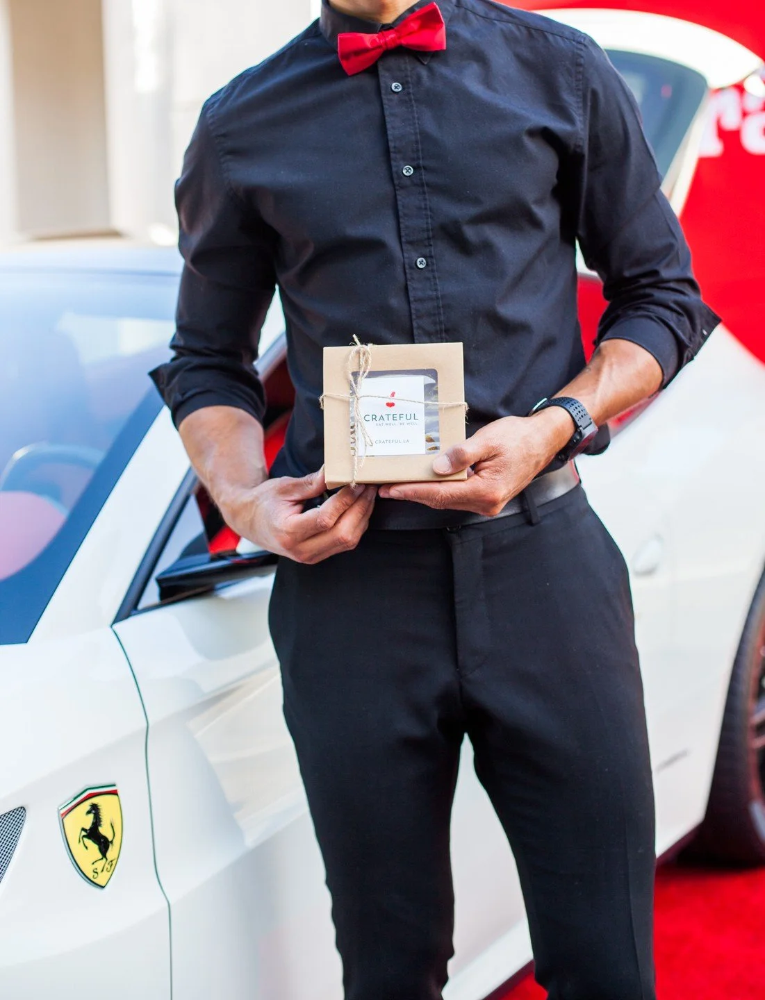 Server in black with a red bow tie holding a Crateful gift box beside a white Ferrari on display