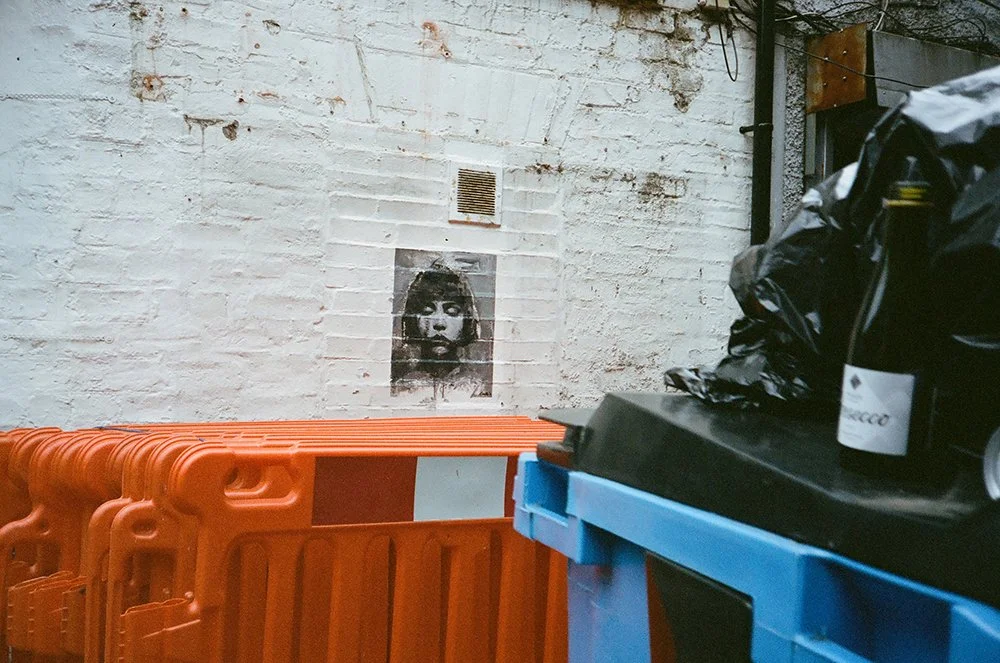 A brick wall with a black-and-white portrait of a person's face with makeup, above a vent. In the foreground, orange barricades and blue plastic containers with black trash bags.