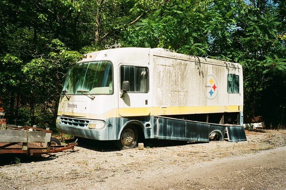 An abandoned food truck with a Pittsburgh Steelers logo, rusted and weathered, parked on a gravel lot surrounded by trees.