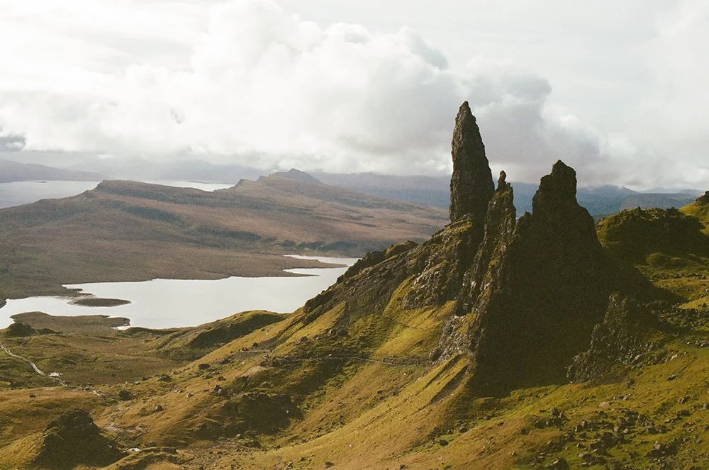 Rugged mountainous landscape with jagged rock formations, rolling grassy hills, a lake, and a cloudy sky.