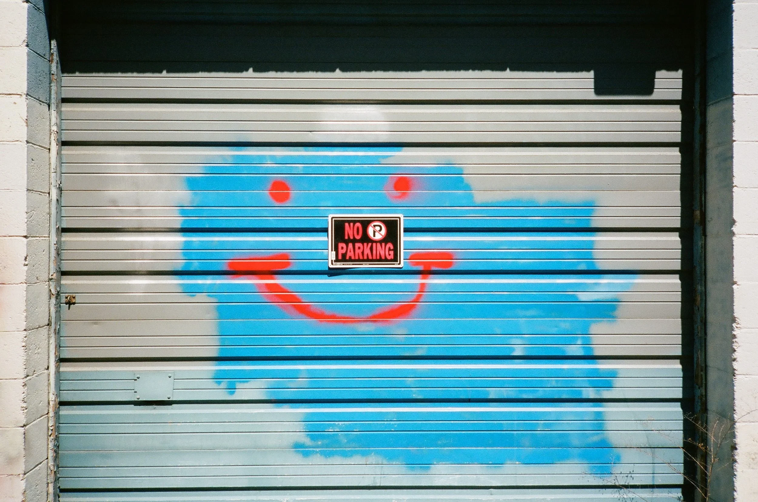 A garage door with graffiti of a blue smiley face, red circles for eyes, and a red smile. There is a black and red "No Parking" sign on the door.