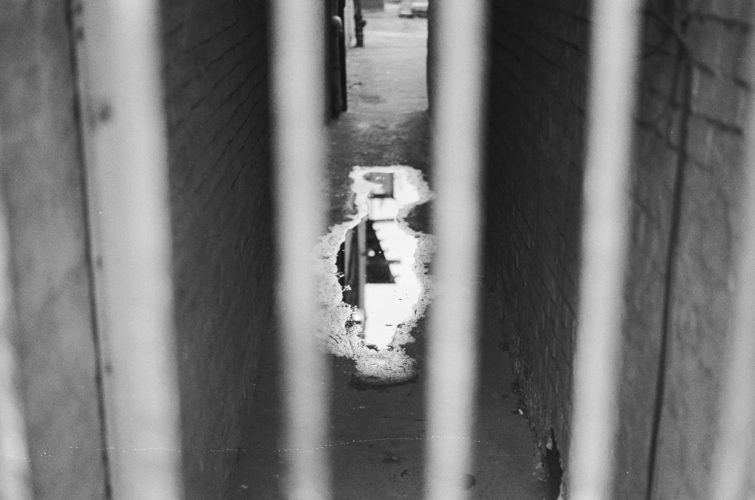 View through metal bars of a narrow alleyway with brick walls on either side. Puddles on the ground reflect the sky and surrounding structures, and the scene appears to be in black and white.