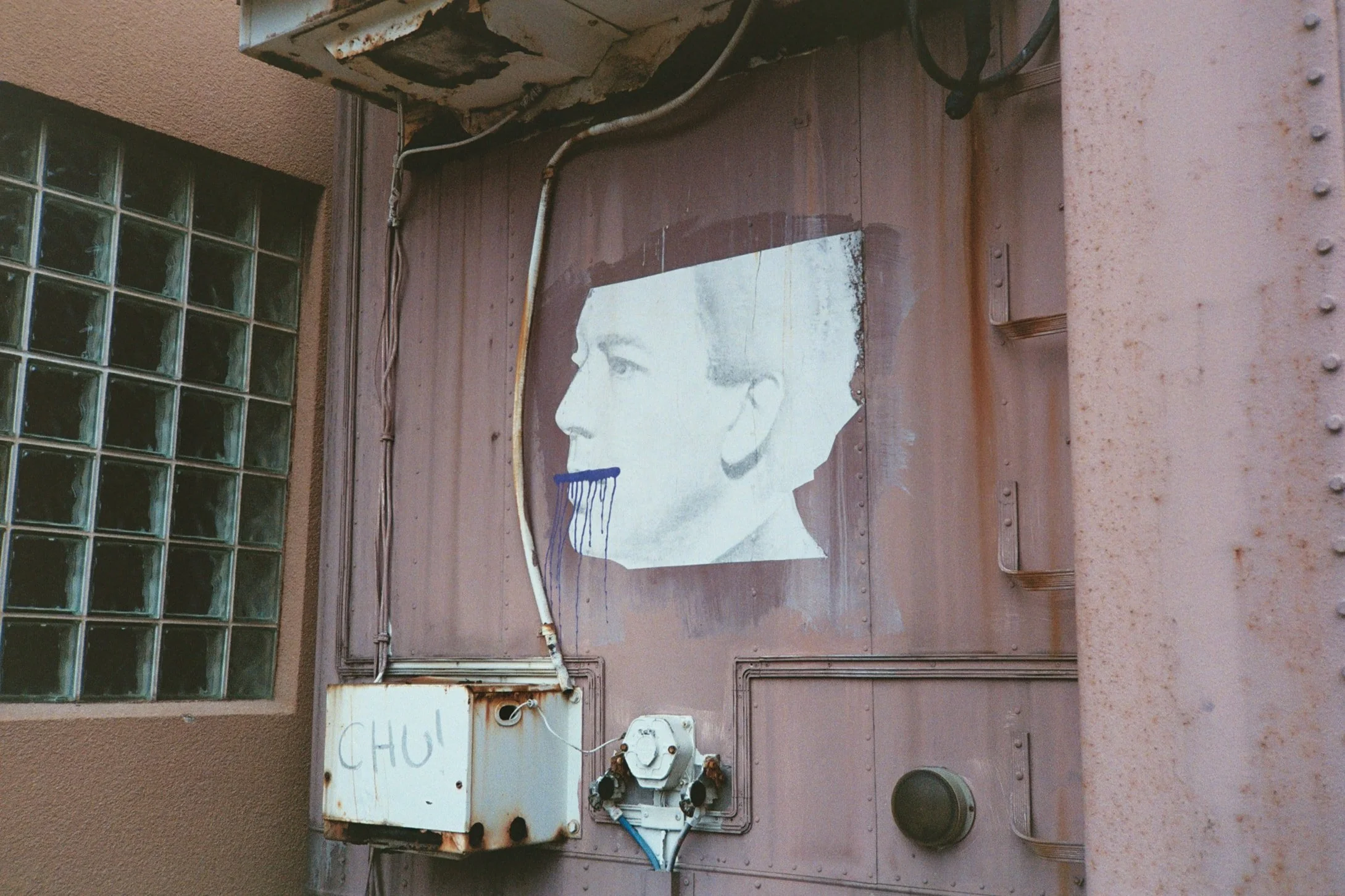 An old, rusty metal outdoor electrical panel box mounted on a wall, with exposed wires and a partially peeled sign that reads 'CHU!'. There is a glass block window to the left and a beige building wall to the right.