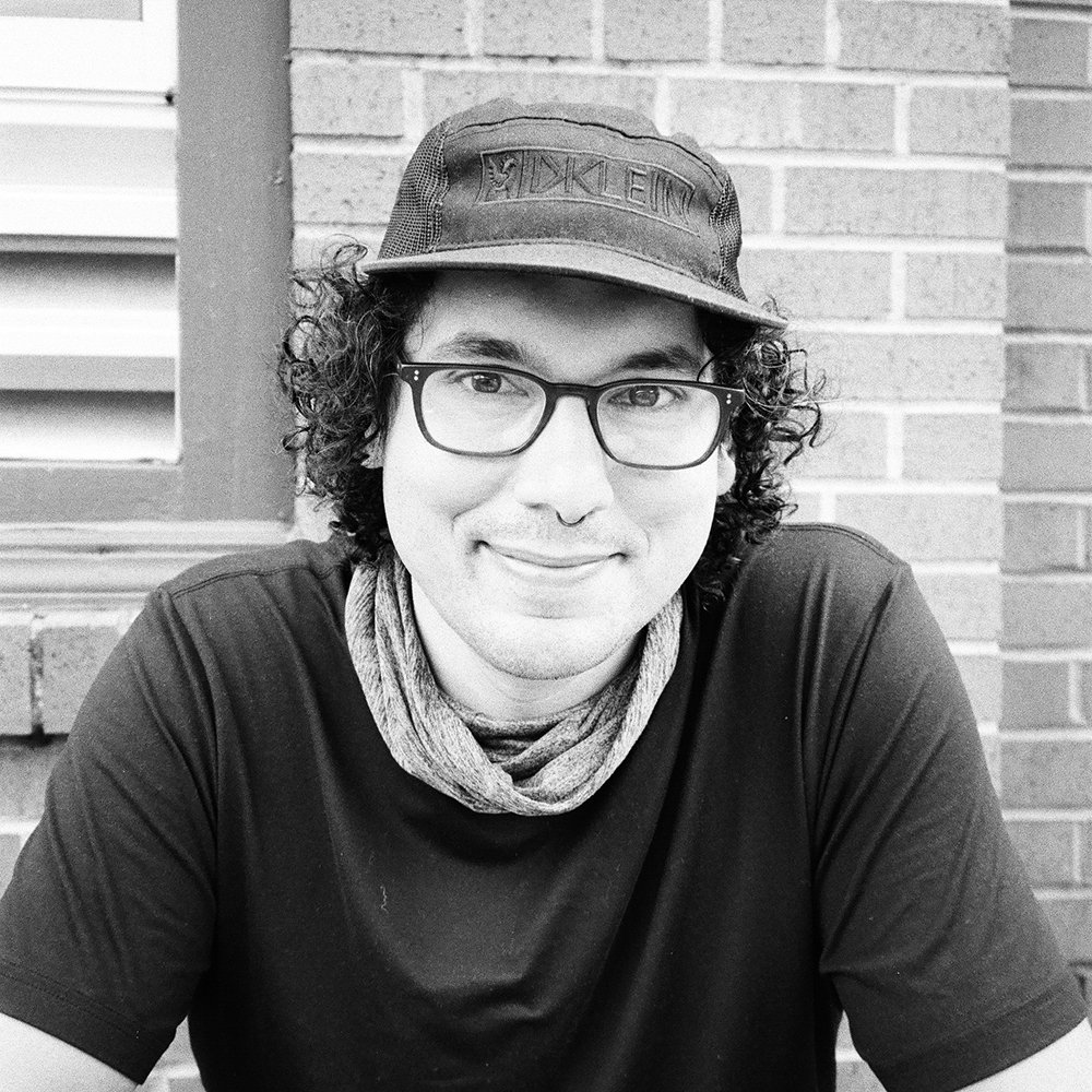 A young man with curly hair, wearing glasses, a cap, a scarf, and a T-shirt, smiling, with a brick wall in the background.