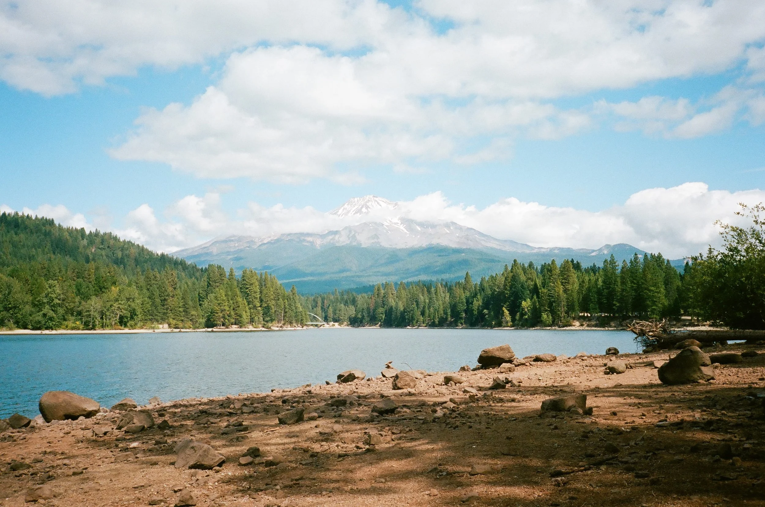 Scenic view of a lake with a forest on the far shore, snow-capped mountains in the background, and a cloudy sky.