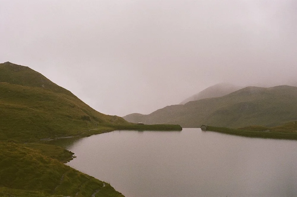 A serene landscape of a large lake surrounded by green rolling hills under a cloudy, foggy sky.