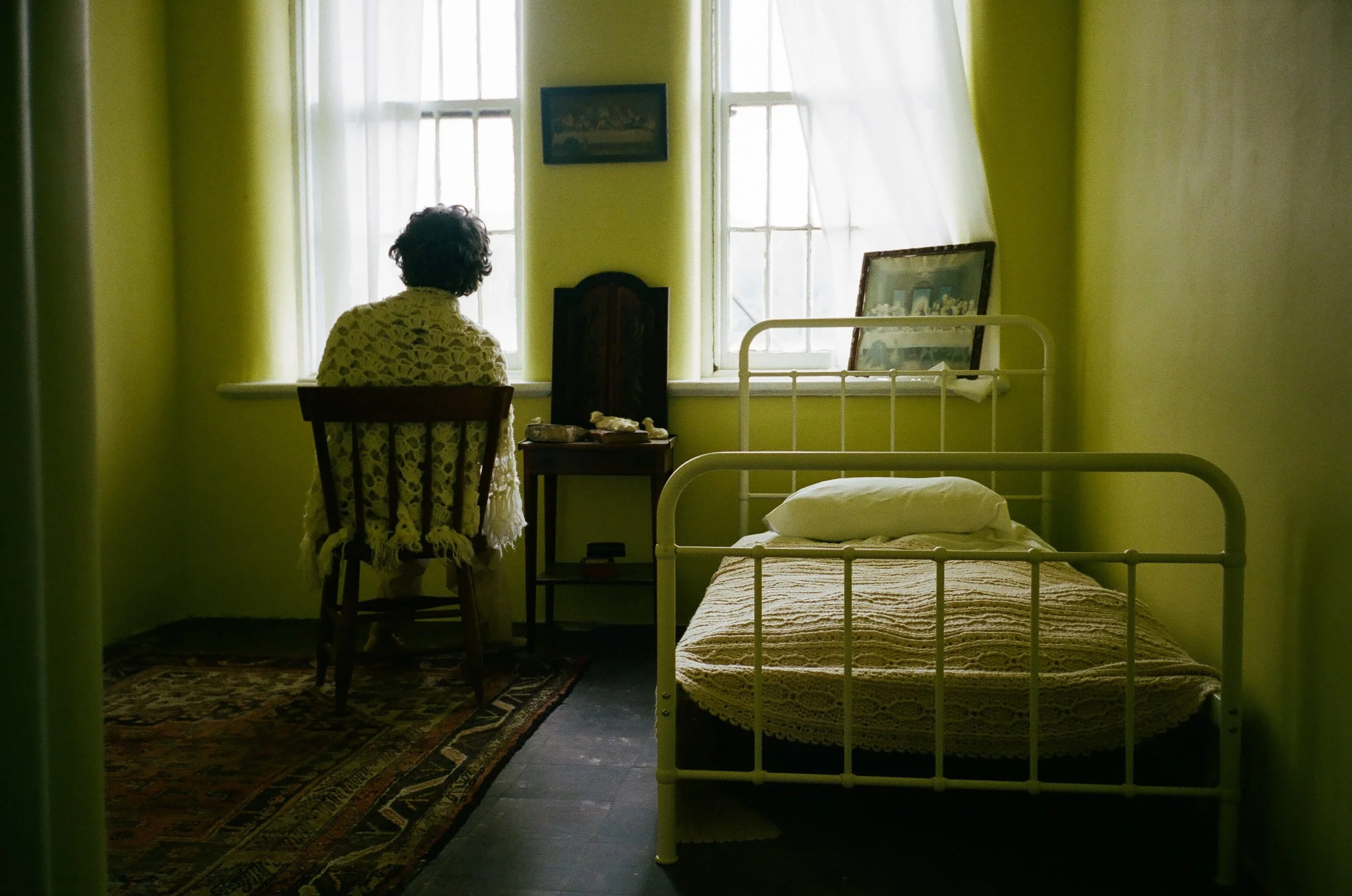 A person with curly hair sitting on a chair facing a window in a room with yellow walls, a bed with a yellow cover, a wooden table with rocks, framed pictures, and paintings on the walls.