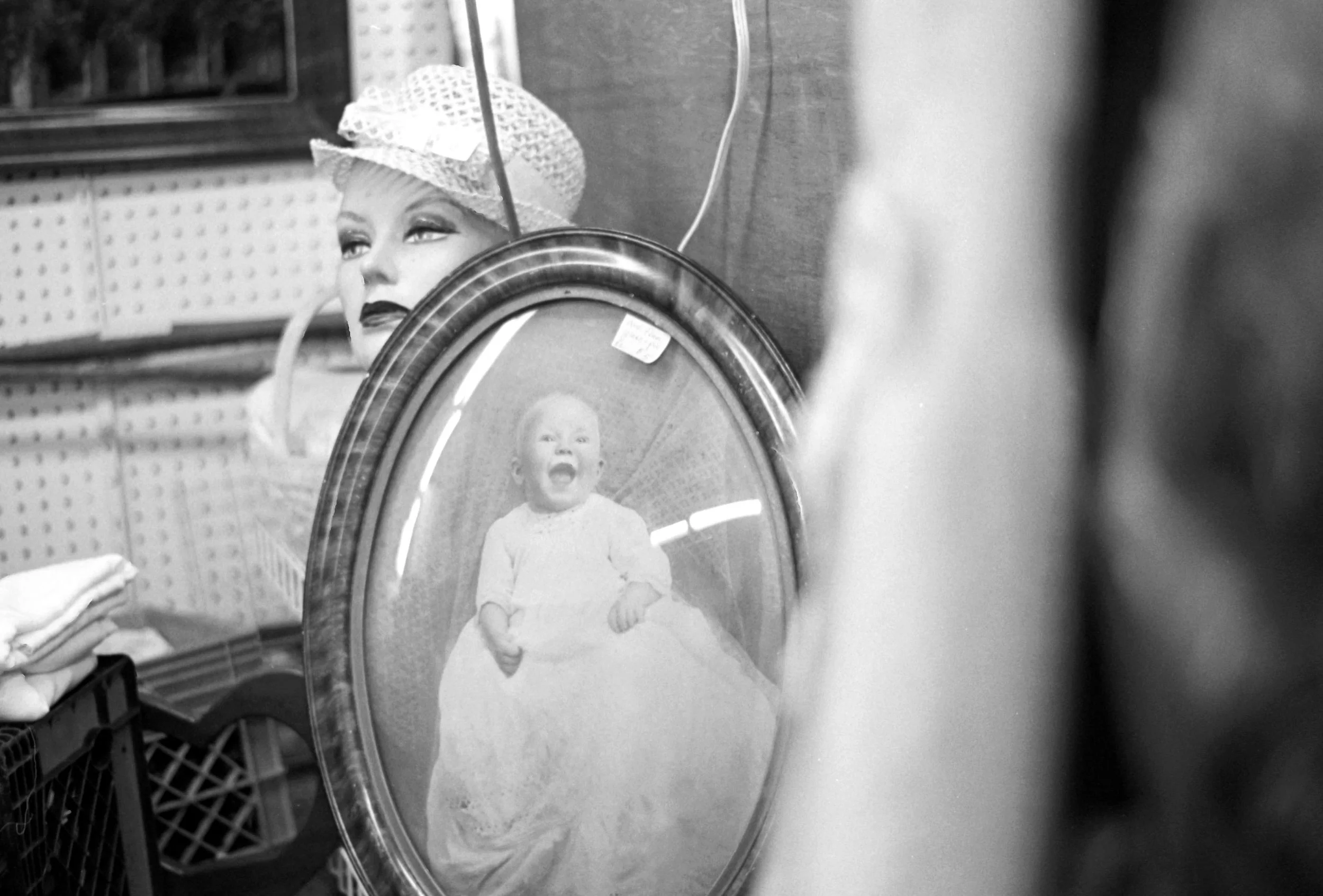 A woman wearing a hat looks at herself in a small round mirror on a store shelf. The mirror reflects a young child sitting, smiling, and looking towards the camera.