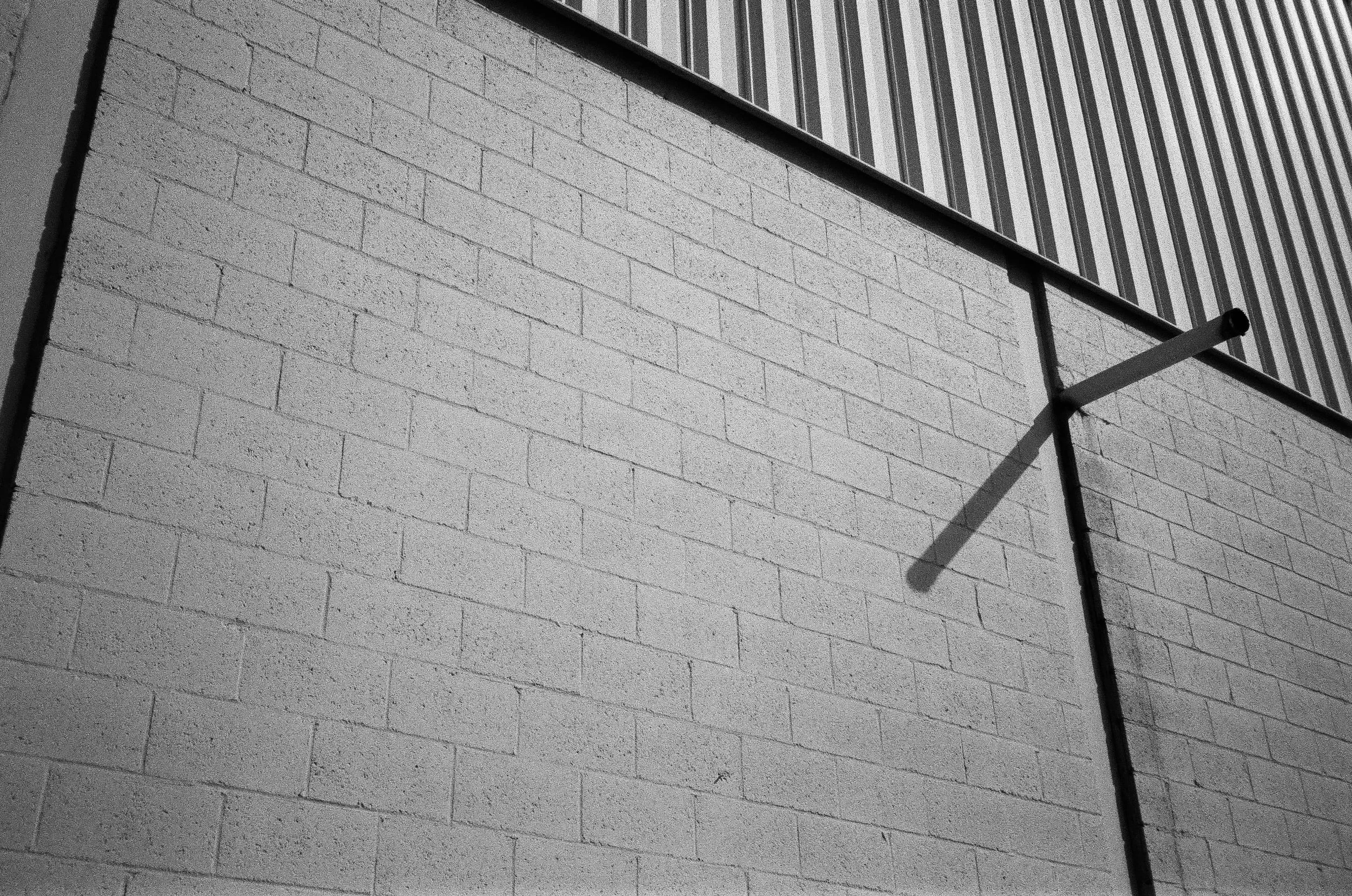 Black and white photograph of an exterior brick wall with a metal pipe extending from it. The wall has a textured surface, and part of a building with vertical siding is visible at the top.
