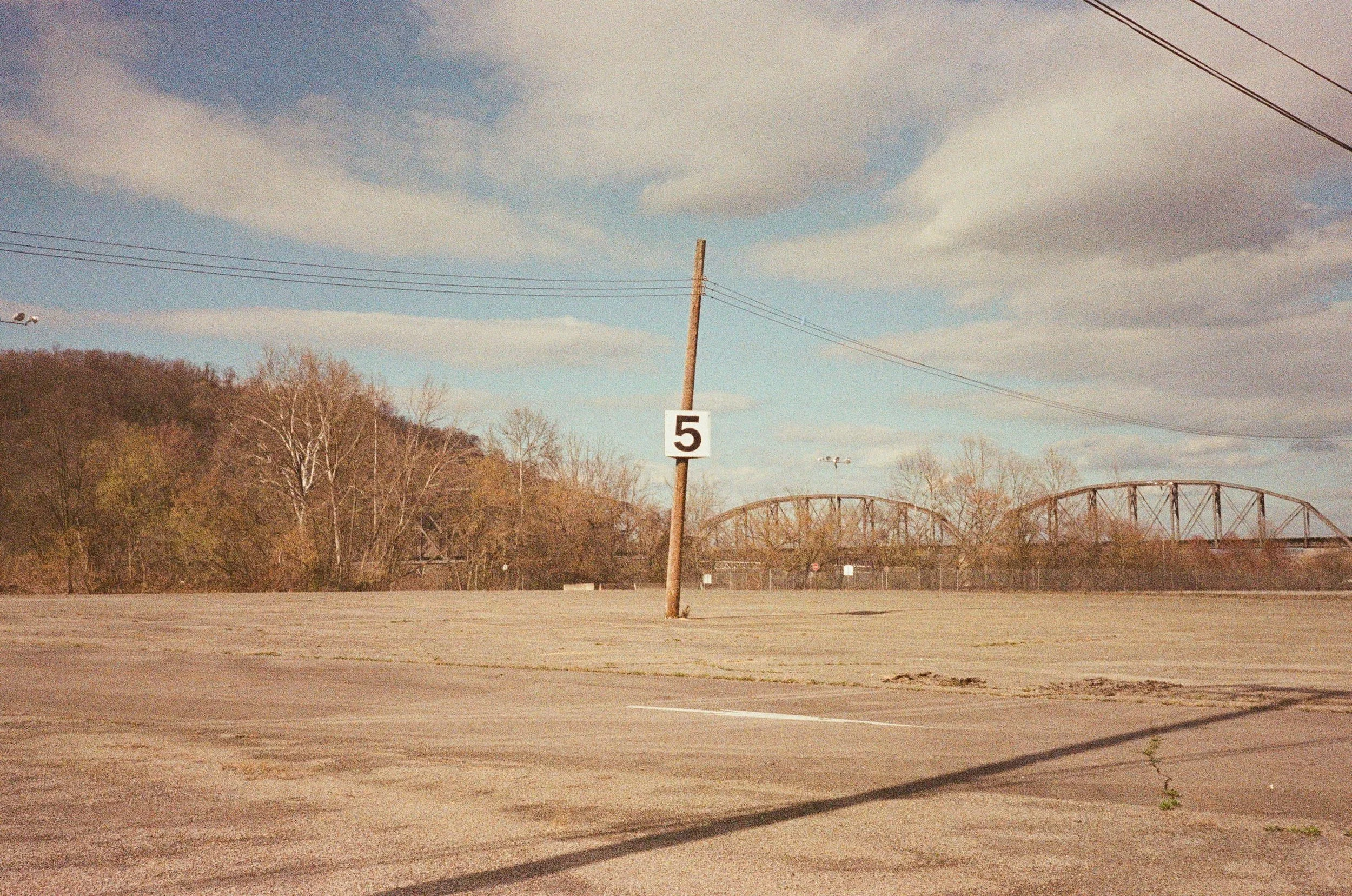 A parking lot with a single wooden utility pole holding a white sign with the number 5. In the background, there are trees with no leaves and a bridge. The sky is partly cloudy.