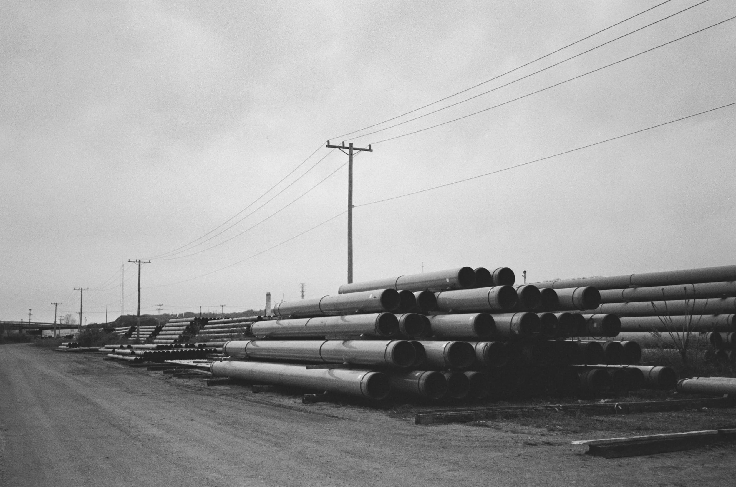 Stacked metal pipes in an industrial yard with power lines and utility poles in the background, shot in black and white.