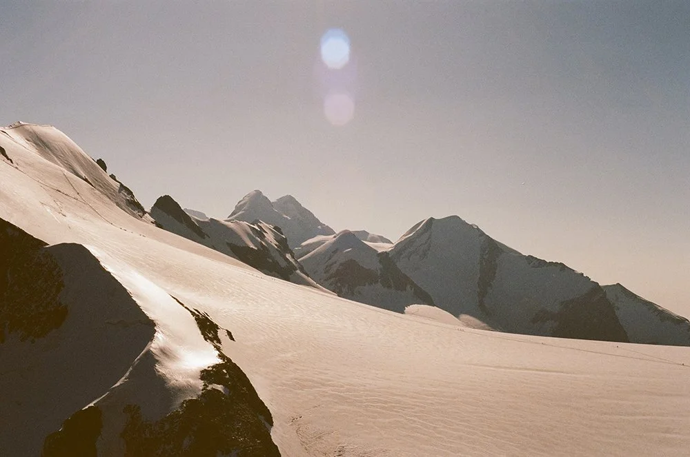 Snow-covered mountain range under a clear sky, with visible lens flare from bright sunlight.