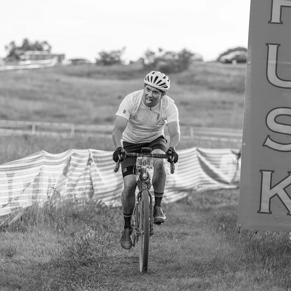 A man wearing a helmet and cycling shorts rides a mountain bike on a grassy trail, expressing effort and concentration.