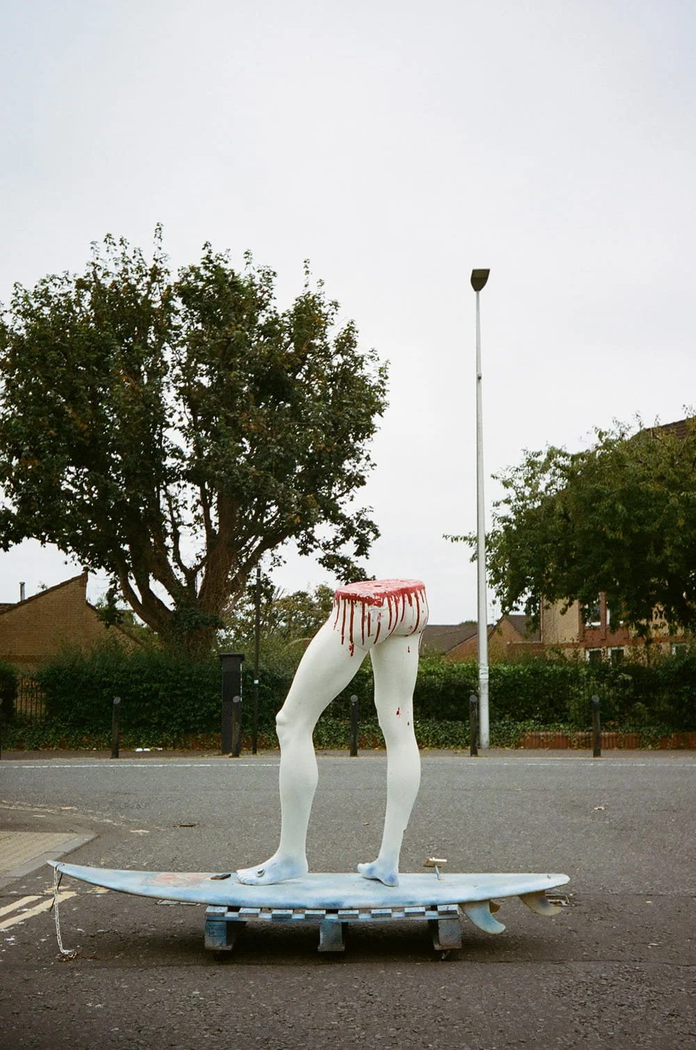 Public art sculpture of a pair of legs with a bloody severed torso on a skateboard in an outdoor setting, with trees, houses, and a lamppost in the background.