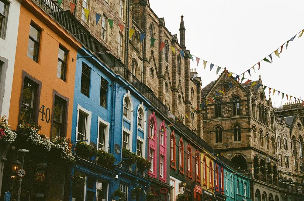 Colorful row of buildings with flower boxes and bunting flags hanging, historic stone church building in the background, overcast sky.