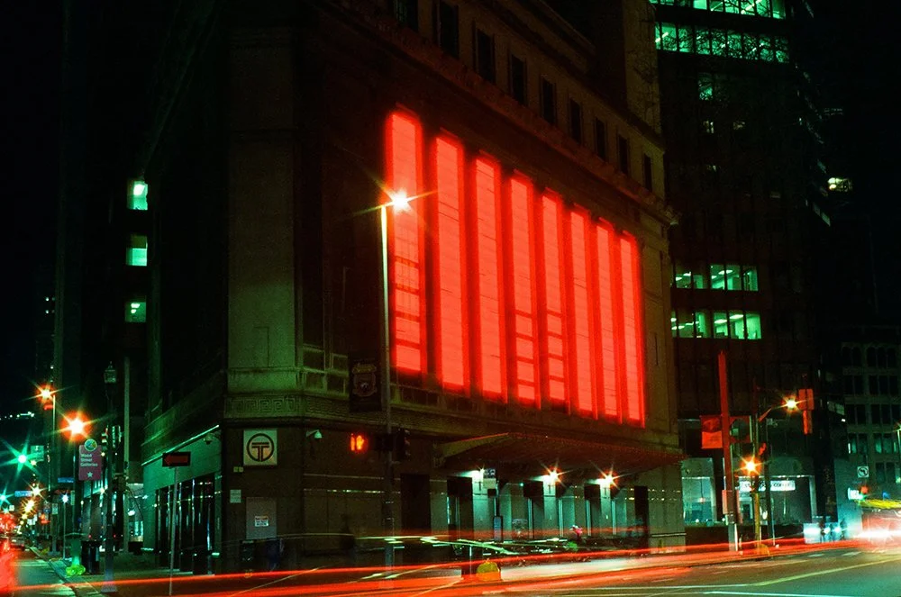 Night urban scene with a building illuminated by large red neon lights on its facade, surrounded by street lights and light trails from moving vehicles.