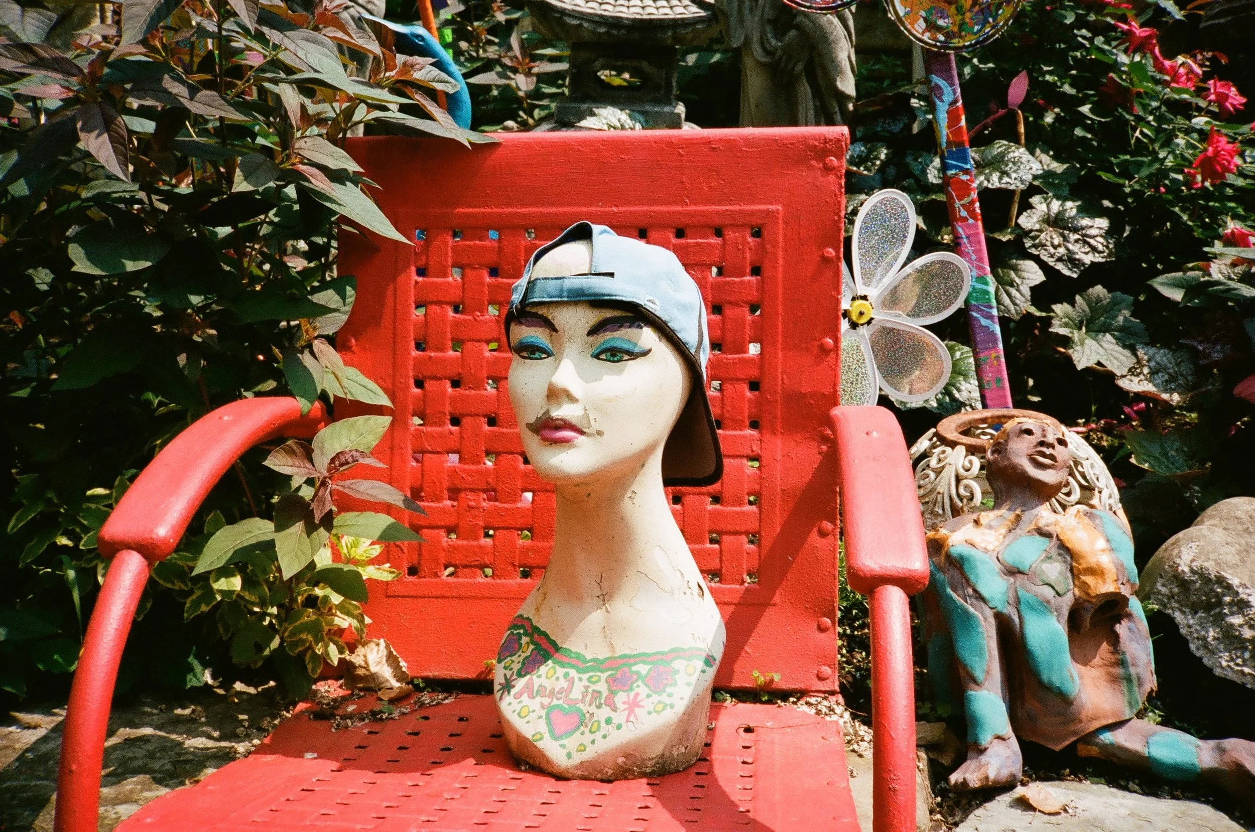 A mannequin head with painted makeup and a white cap, placed on a red chair outdoors among plants and decorative objects, including a butterfly ornament and a colorful rod.