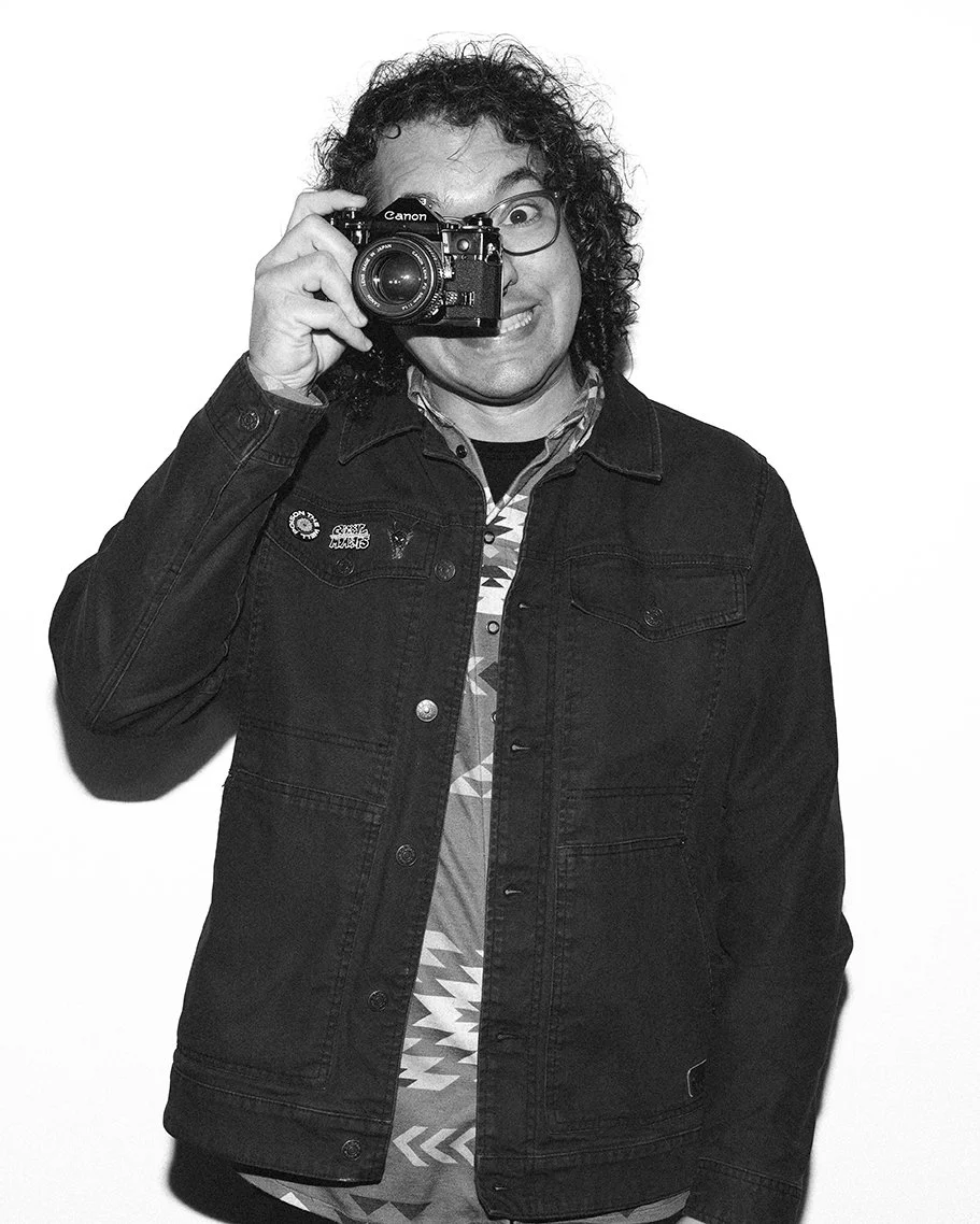 A person with curly hair and glasses holding a camera in front of their face, smiling, in front of a plain background.