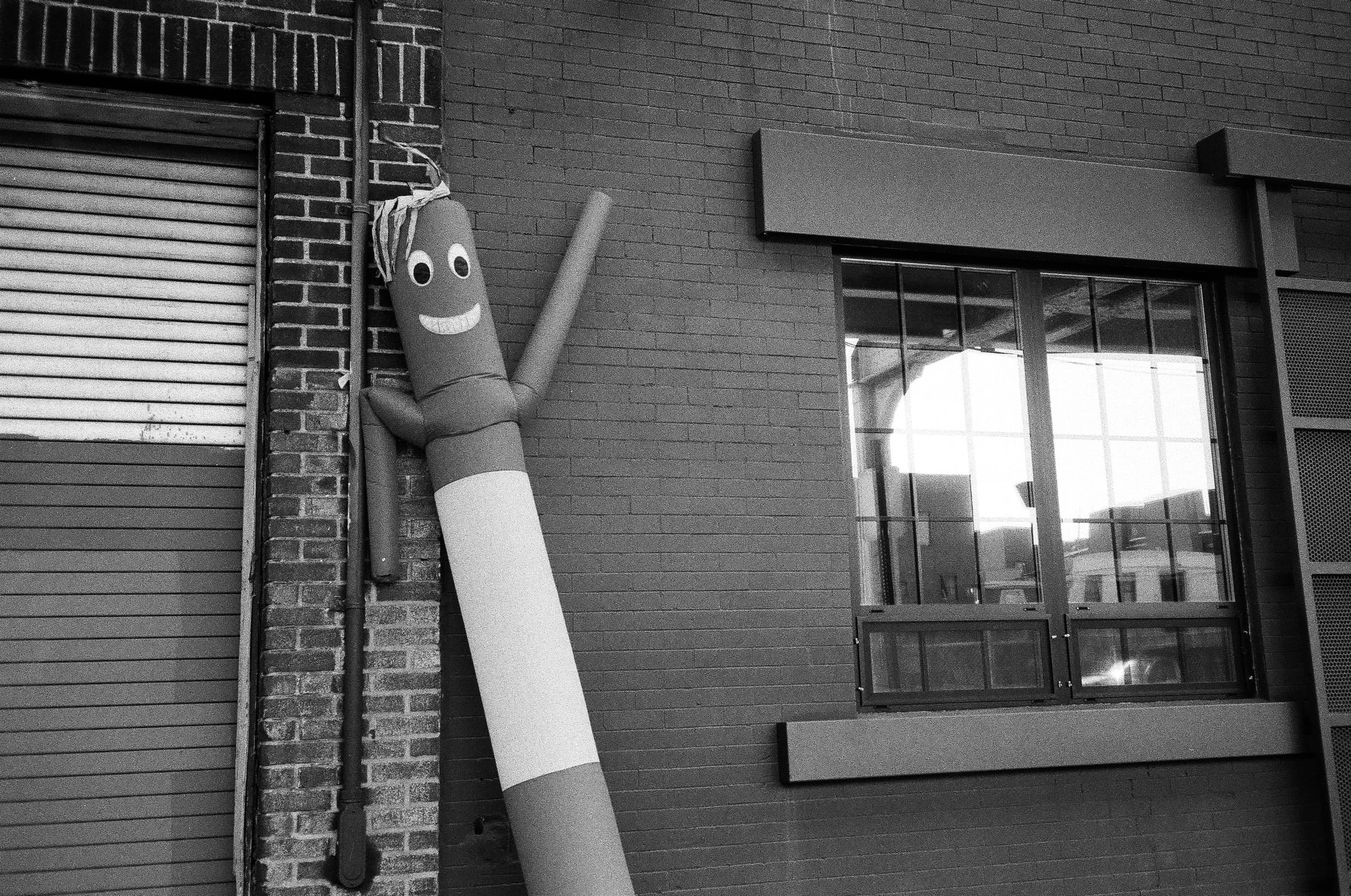 A sidewalk scene featuring a large inflatable tube man with a smiling face, leaning against a brick wall and a large window with a metal frame, on an urban building exterior.