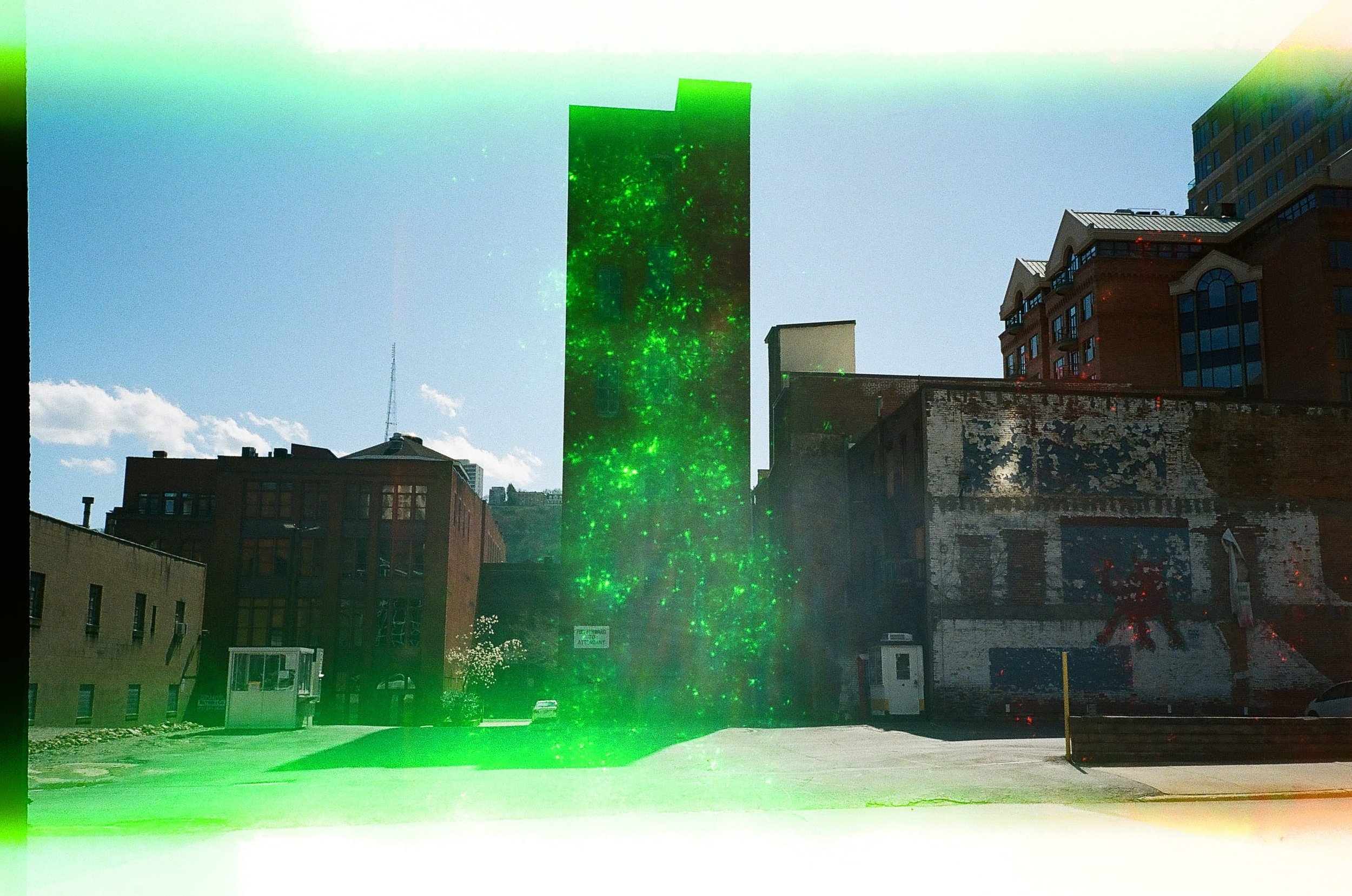 Cityscape with a tall building emitting green laser or light beams, other buildings, and a parking lot with some cars.