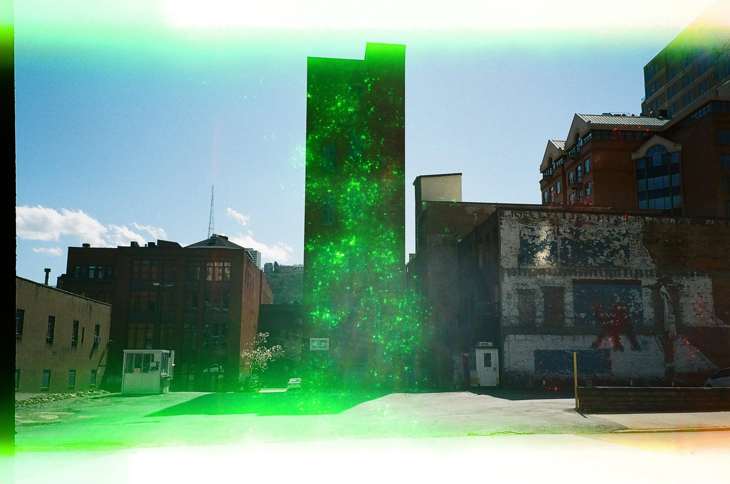 Cityscape with a tall building emitting green laser or light beams, other buildings, and a parking lot with some cars.