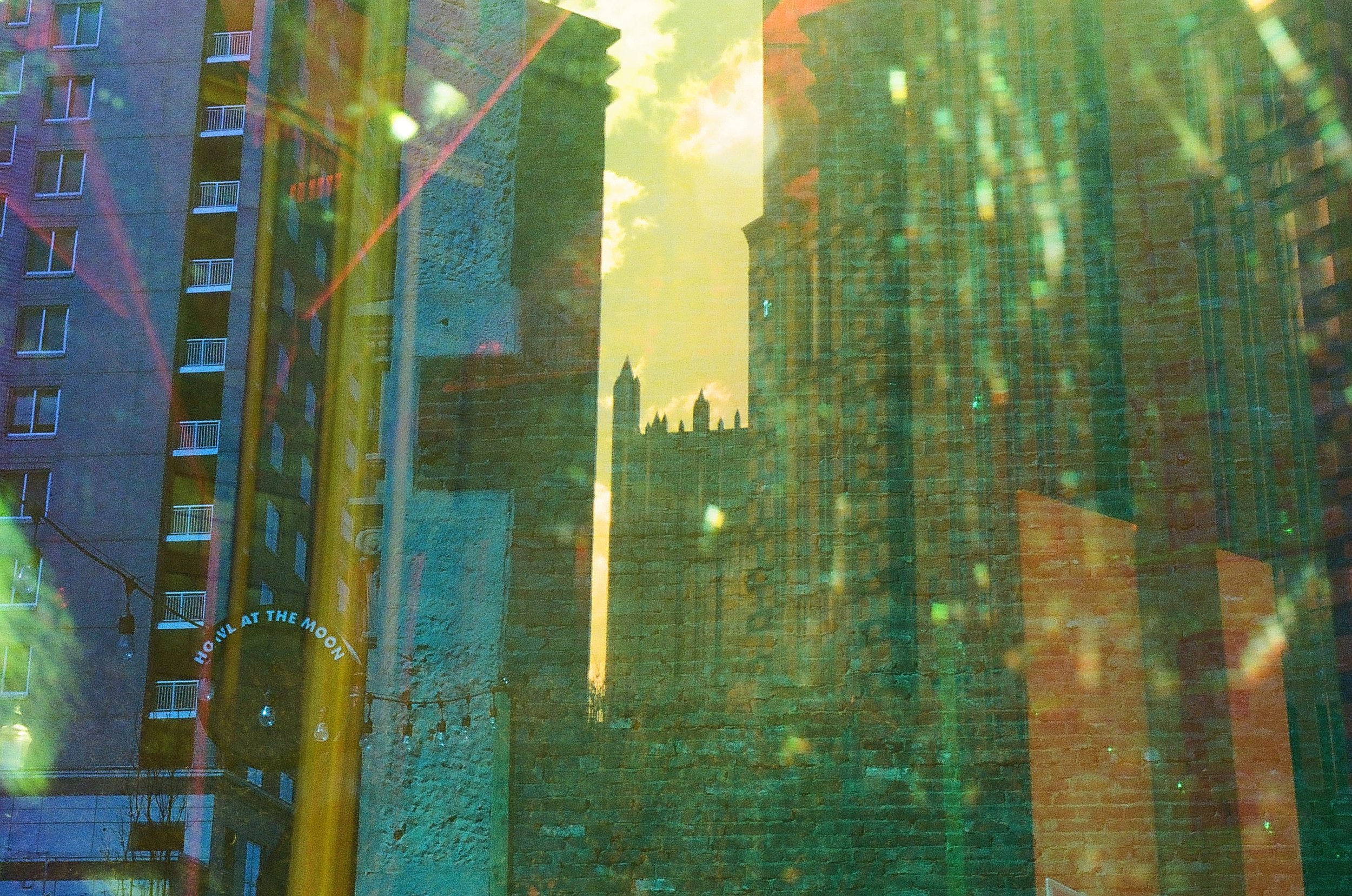 A city scene with buildings, a sign that reads 'HOP ON, HOP OFF AT THE MOON,' and the silhouette of tall structures in the distance, with sunlight and cloudy sky overhead, featuring colorful reflections.