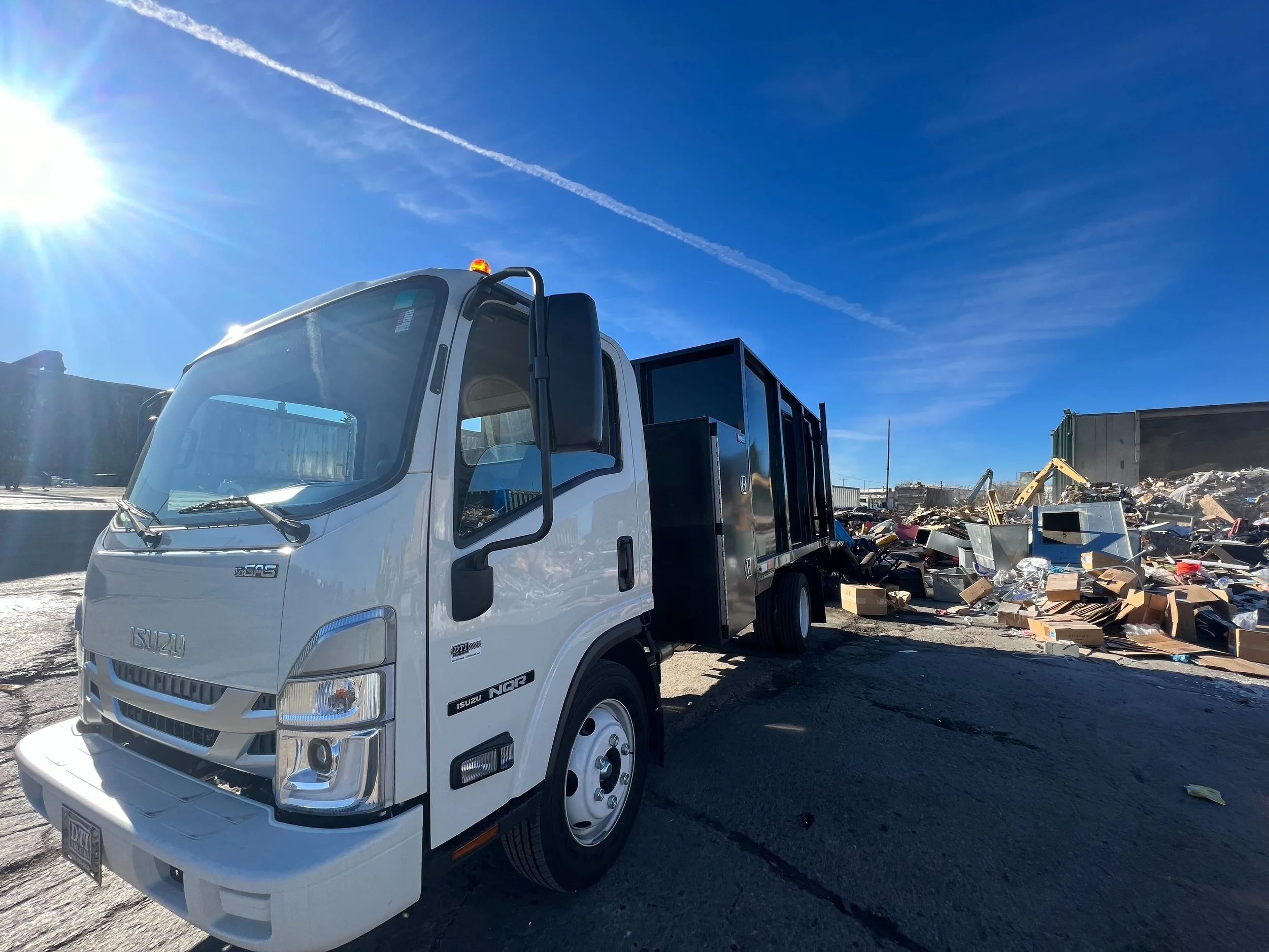 White Isuzu garbage truck with black open cargo area parked on a paved surface, with a pile of trash and debris in the background under a sunny blue sky.