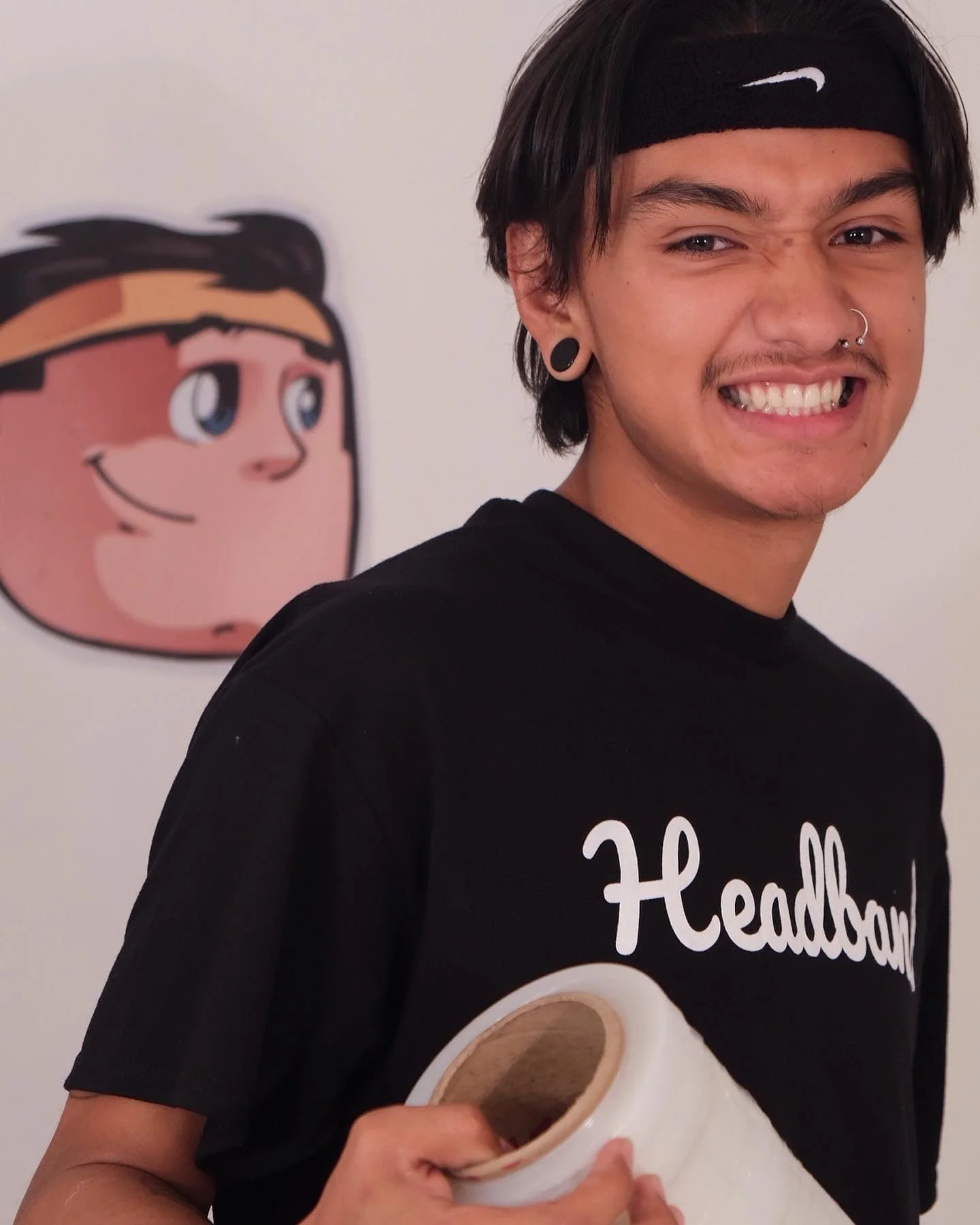 A young man with dark hair, multiple piercings, and stretched earlobes, smiling with gritted teeth, wearing a black t-shirt with white text and a black Nike headband, holding a roll of masking tape, standing in front of a cartoon character image on a white wall.