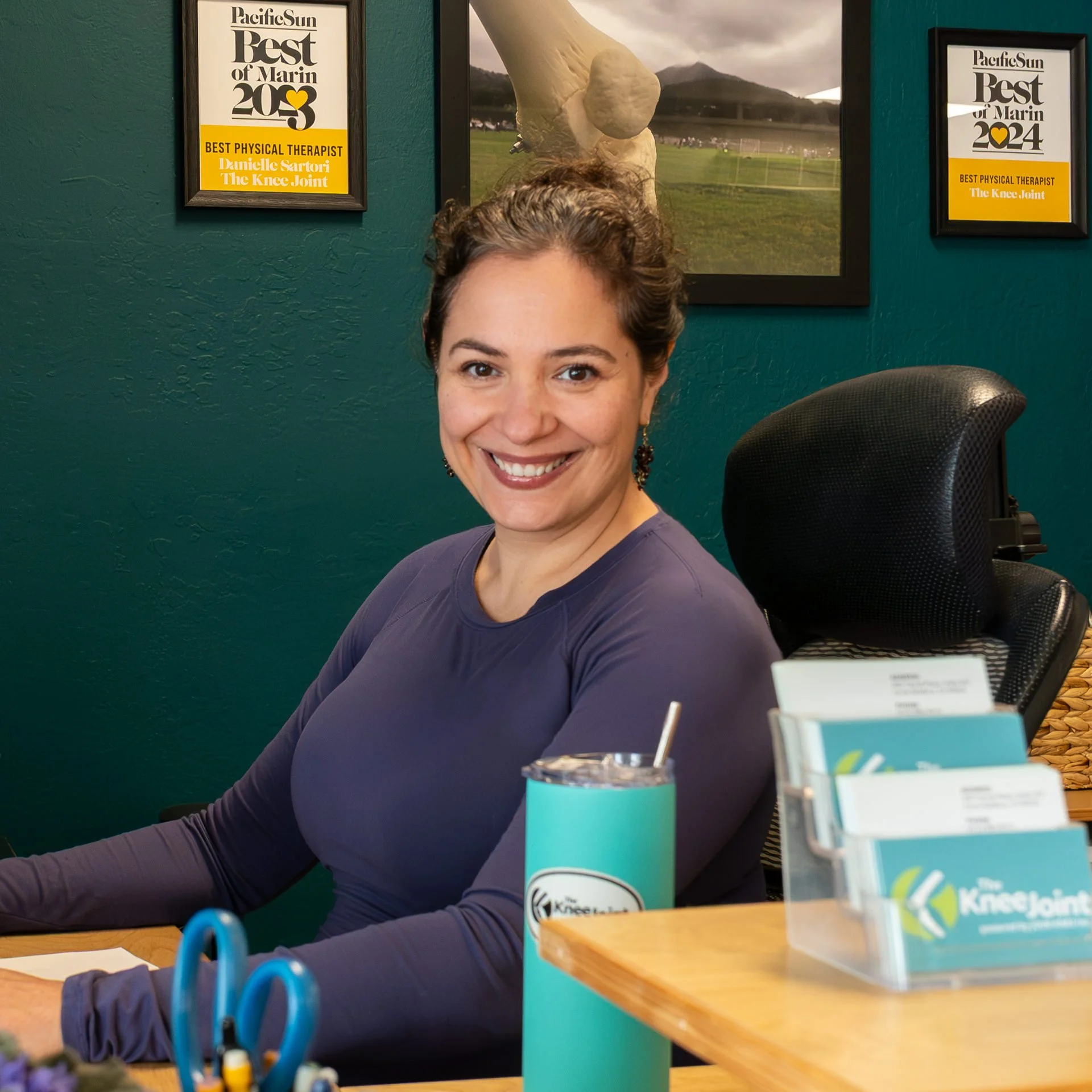 Julia Zave with dark curly hair smiles at a front desk in front of a teal green wall displaying three framed Pacific Sun Best of Marin awards (2023 - 2025) for Best Physical Therapist, Danielle Sartori of The Knee Joint.