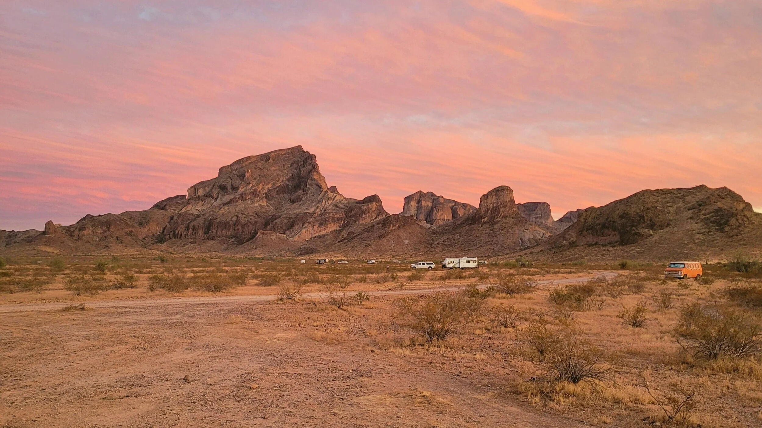 Desert landscape with rocky mountains, sunset sky, and parked vehicles, including a van and a trailer.
