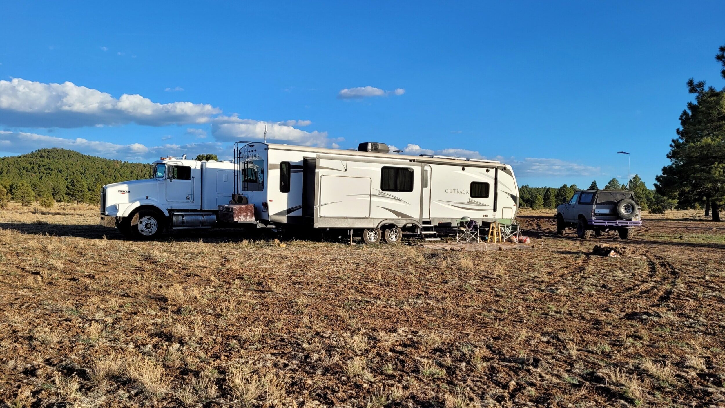 Semi truck and RV in a grassy open field with a pickup truck nearby, surrounded by trees and a clear blue sky.