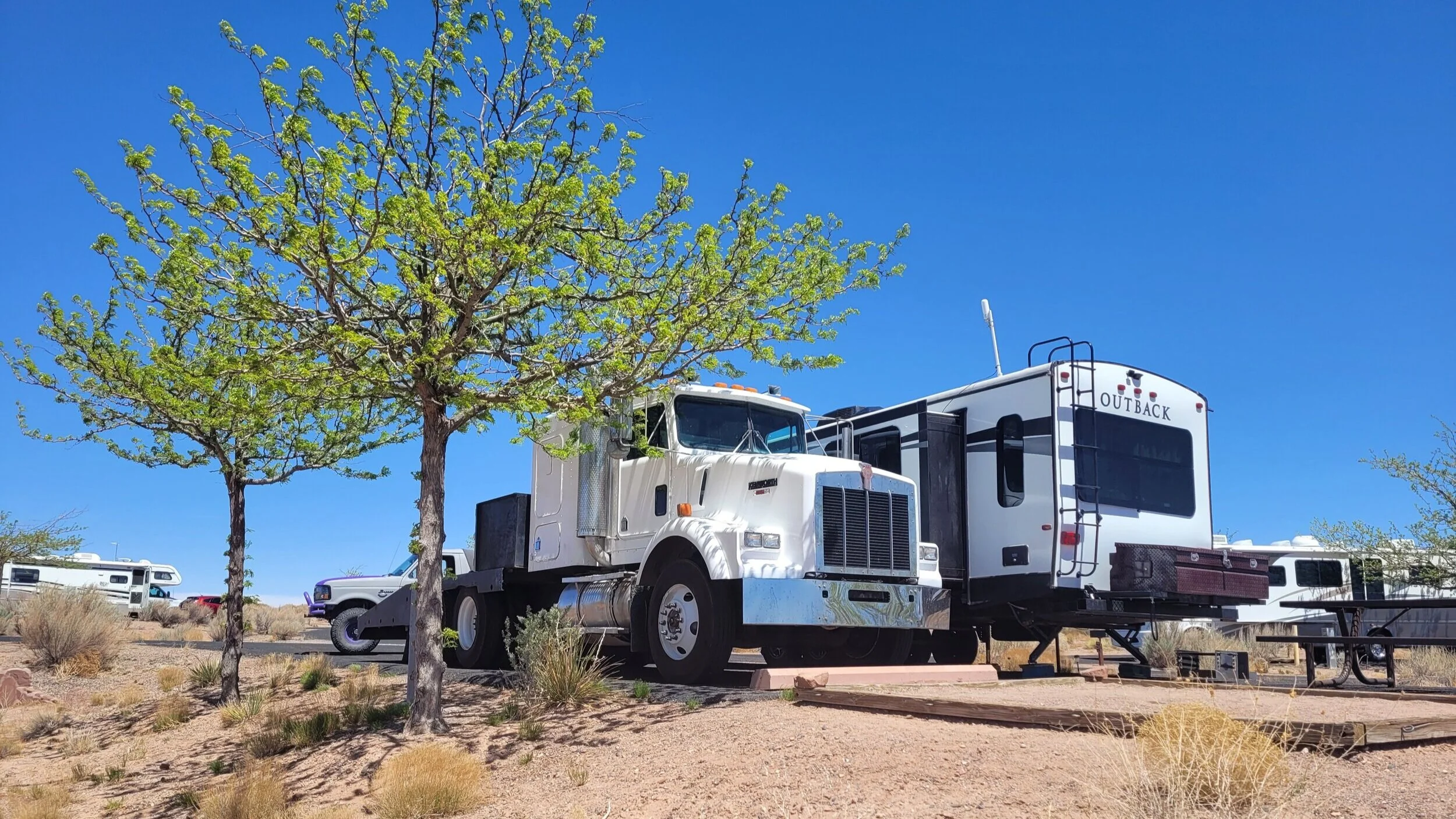 White semi-truck parked next to a large RV camper in a desert campground with clear blue sky and sparse trees.