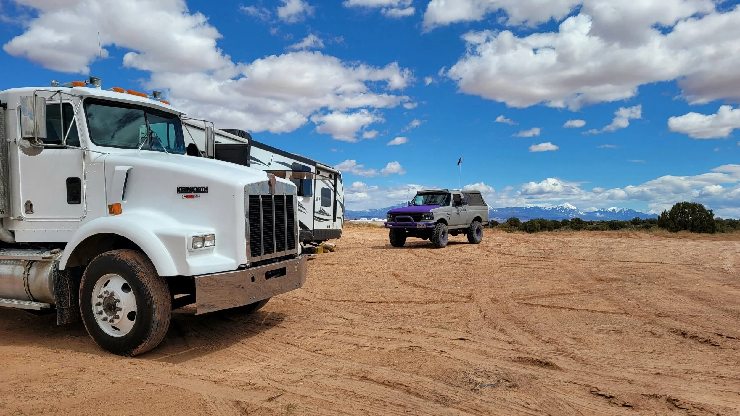 White semi truck, recreational vehicle, and pickup truck parked on a dirt field with mountains and cloudy blue sky in the background.
