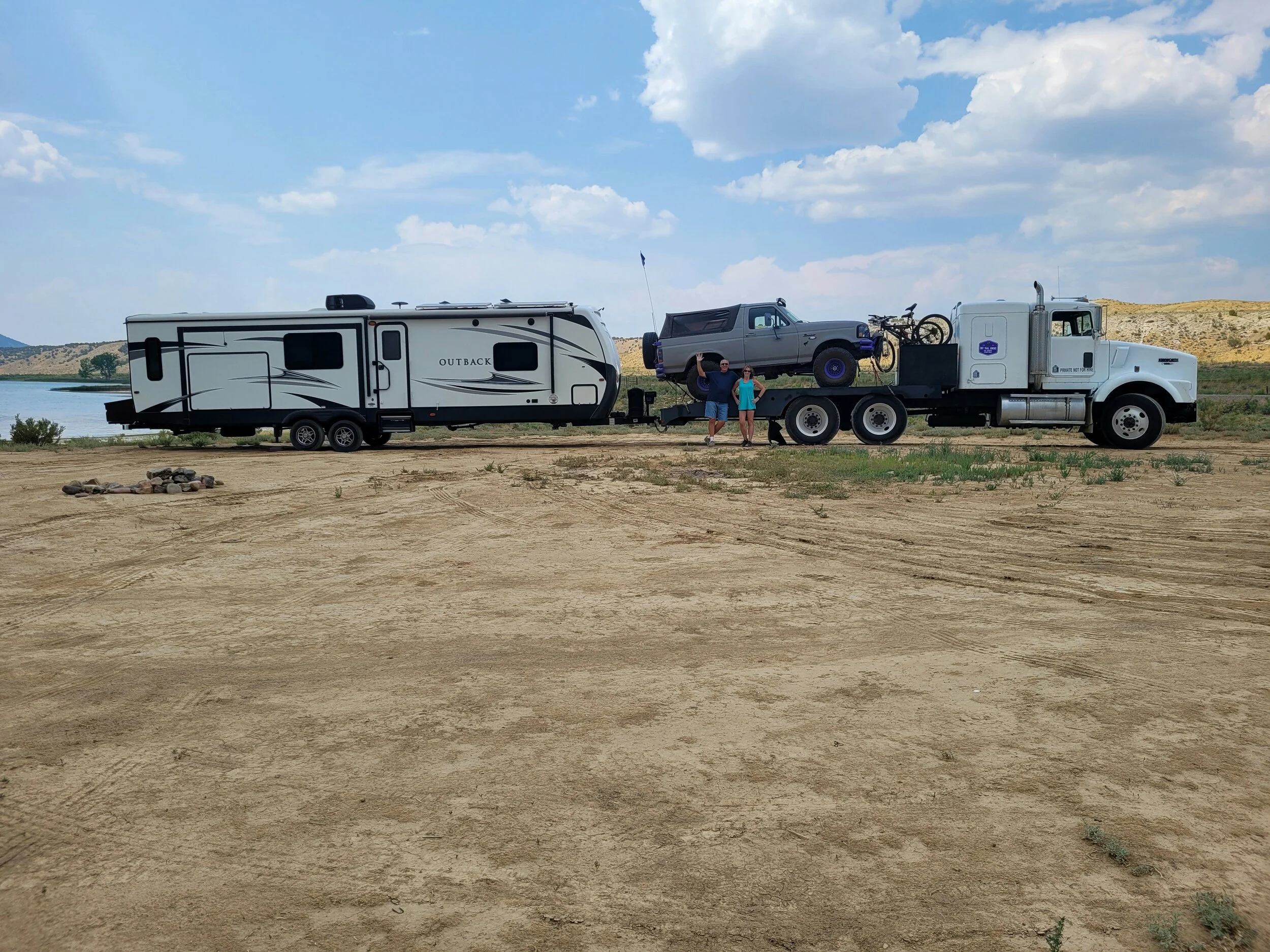 Large fifth-wheel RV trailer parked near a lake, hitched to a flatbed truck carrying a small off-road vehicle and two bicycles, with two people standing nearby.