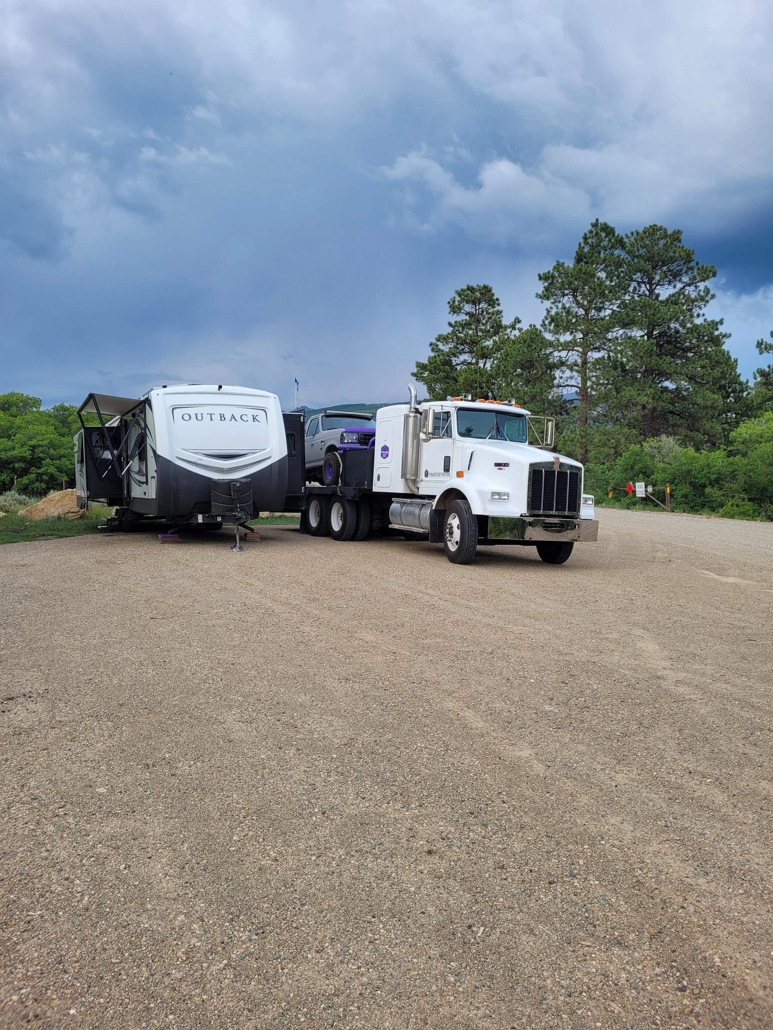 Semi-truck towing a large RV and carrying a Jeep, parked on gravel area under cloudy sky, surrounded by trees.