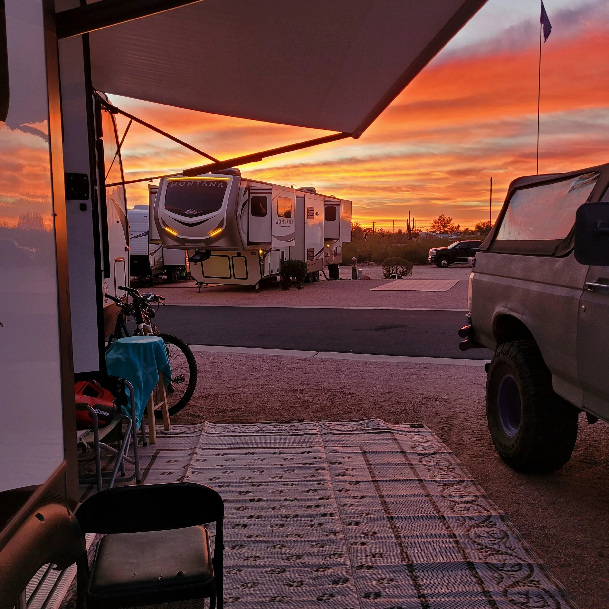 RV parked at a campsite during sunset with a bike, chair, and vehicle nearby.