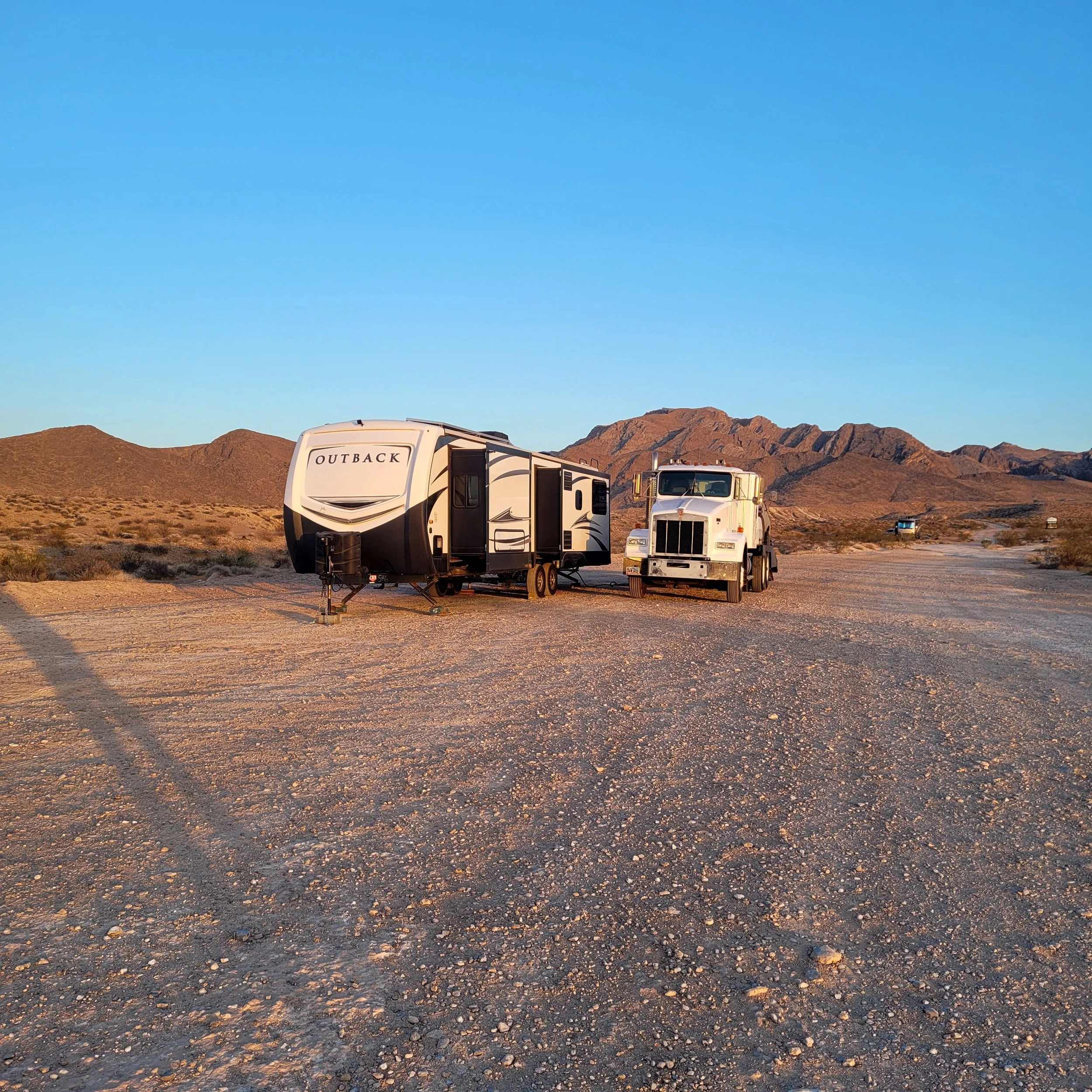 A camper trailer labeled "Outback" parked on a dirt road next to a large white truck in a desert landscape with mountains in the background.