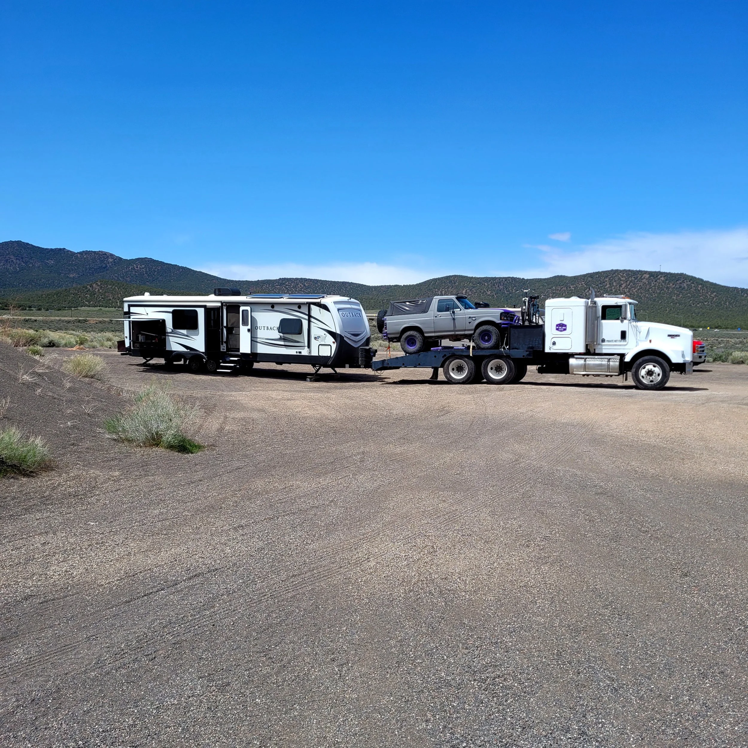 A white semi-truck hauling a travel trailer and a flatbed trailer with an off-road vehicle in a desert setting with mountains in the background.
