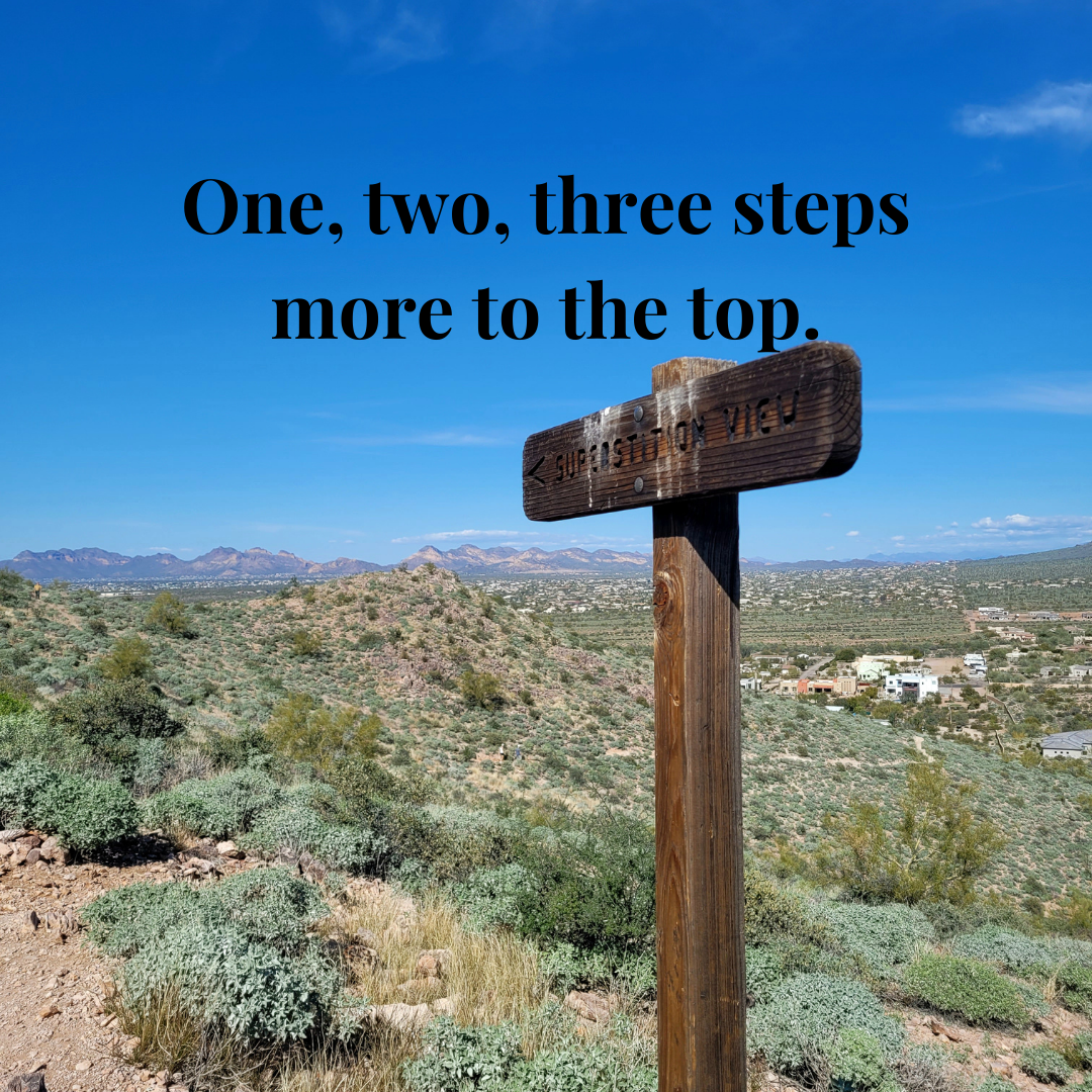 Wooden trail sign labeled "Superstition View" on a mountainous path with a scenic desert landscape in the background and clear blue sky. Text overlay reads "One, two, three steps more to the top."