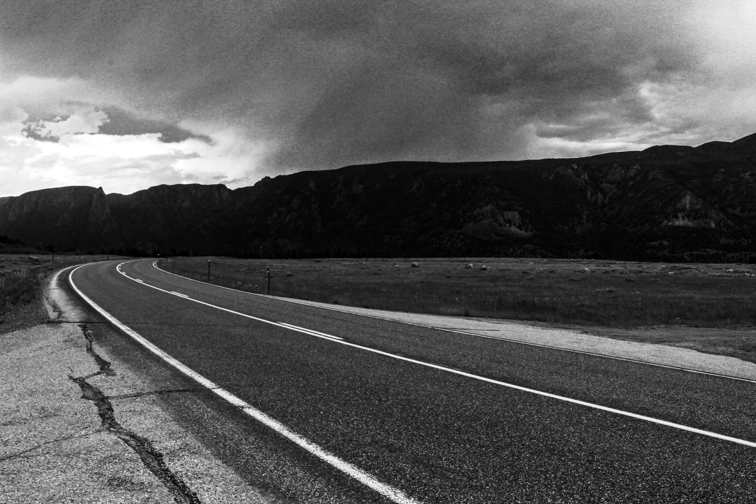 Black and white image of a winding road through a mountainous landscape under a cloudy sky.