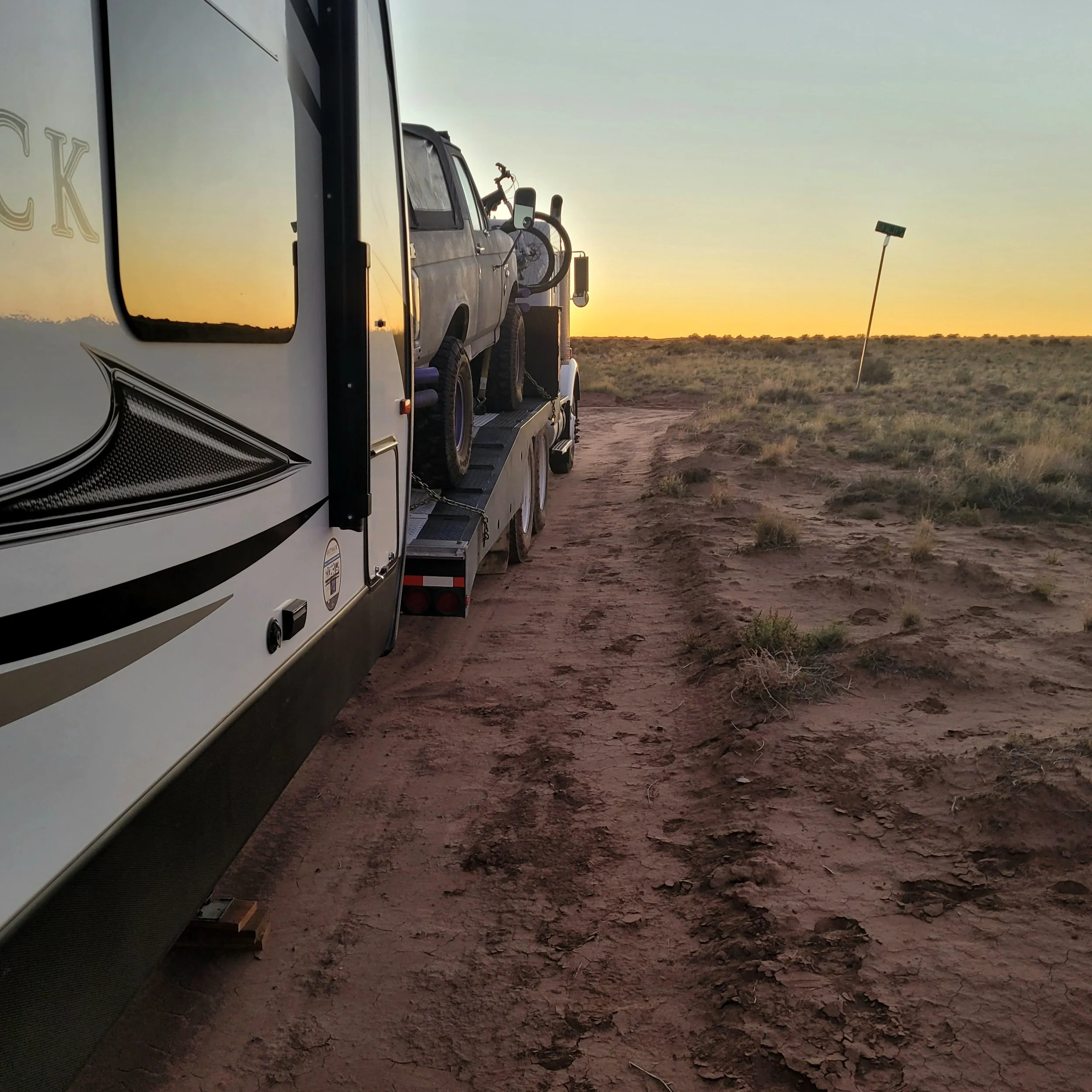 Truck towing a trailer with a vehicle on a dirt road at sunset