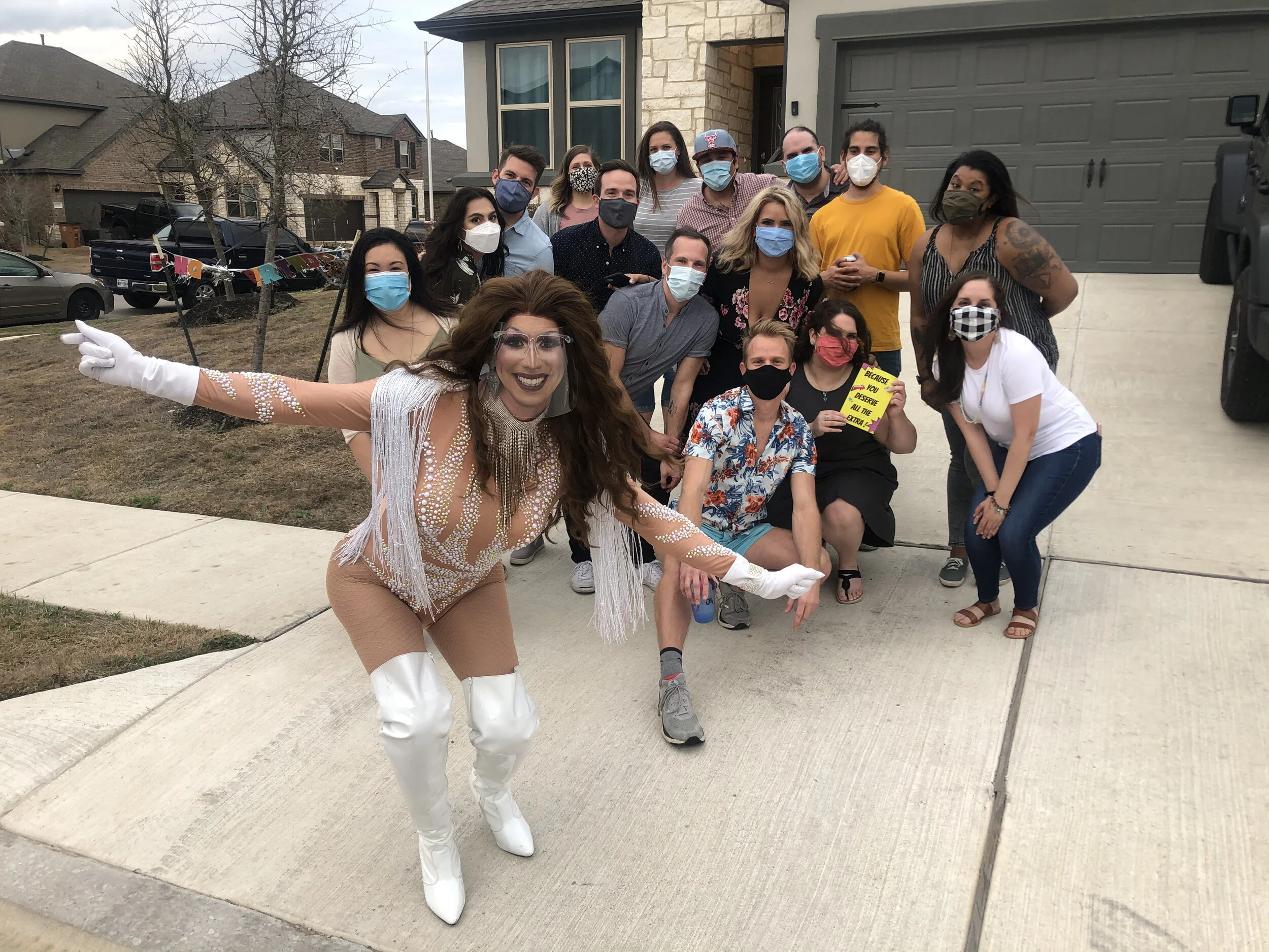 Group of people posing outdoors in front of a house, with a person dressed in a flashy costume standing in front. Many are wearing face masks.