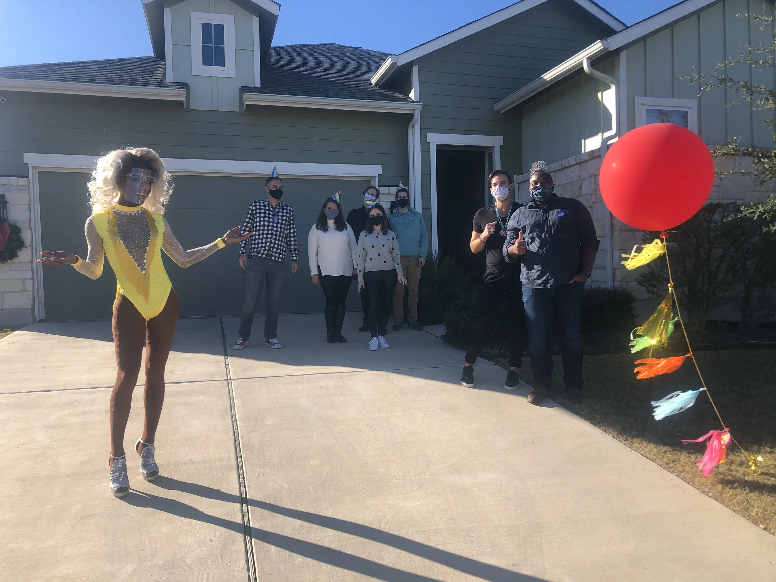 A group of people standing outside a house during a celebration, with a drag queen in a yellow costume and high heels in the foreground, and a large red balloon with colorful tassels attached to it on the right.
