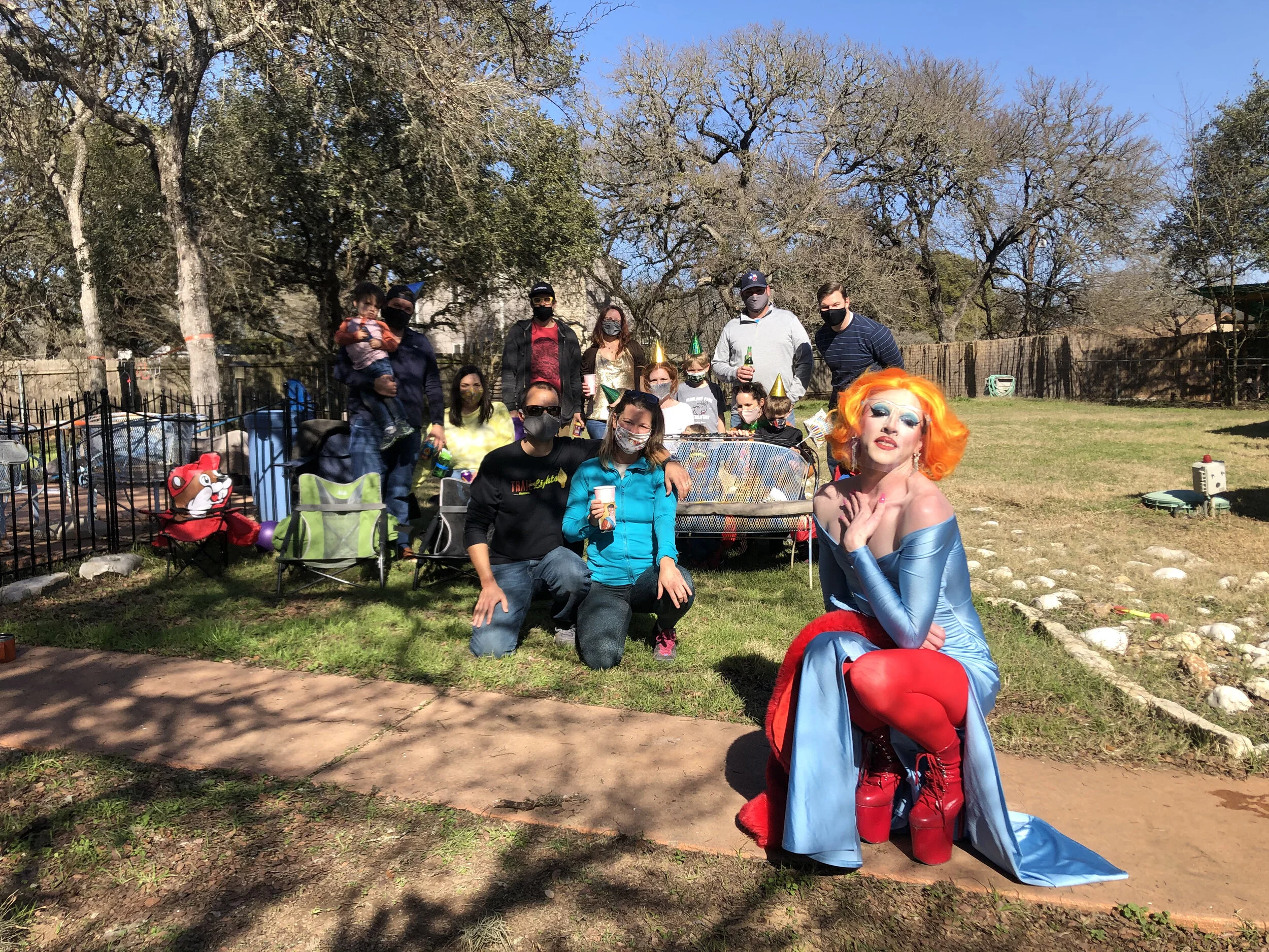 Group of people gathered outdoors in a backyard, celebrating with a person dressed in a colorful drag queen costume with orange hair, blue dress, and red boots. Some are wearing face masks, and a few children are present. There are trees, some with b