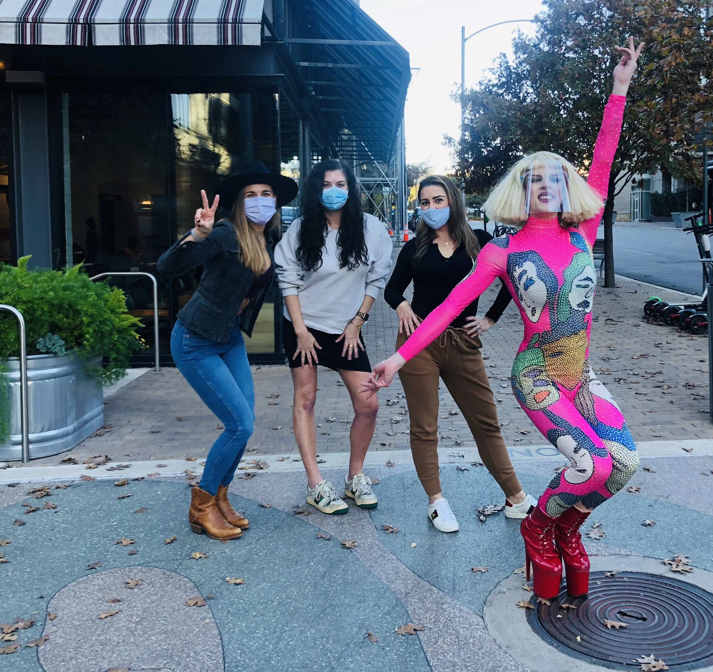 Four women, three wearing face masks, stand and pose on a city sidewalk. The woman on the far right is dressed in a colorful, form-fitting jumpsuit with a pop art design, a face shield, and extravagant red platform shoes. The other women are casually