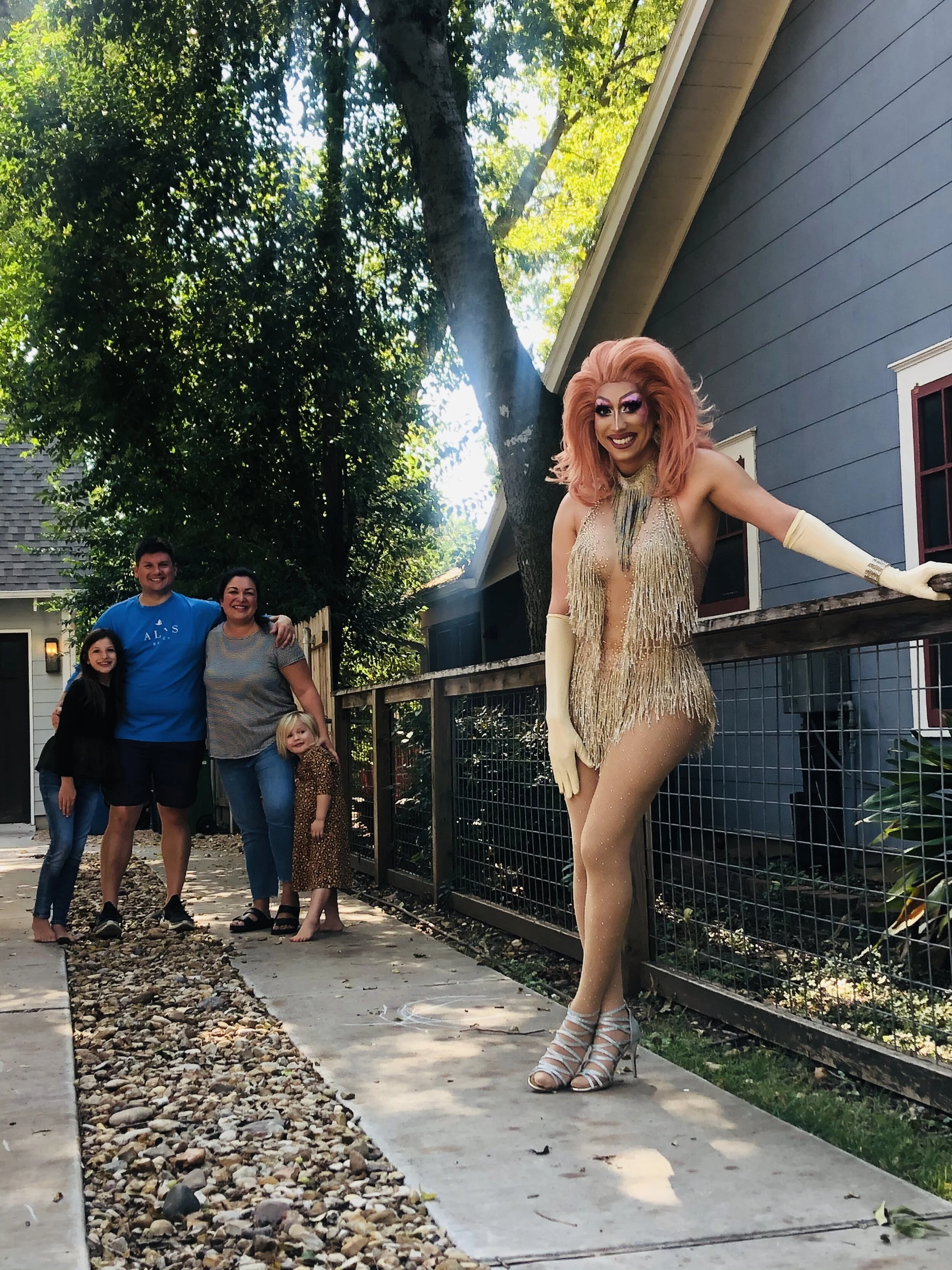 A woman dressed as a drag queen with pink hair, gold fringe dress, and high heels standing on a sidewalk, smiling. In the background, four children are standing together, smiling, near a blue house with trees and sunlight overhead.