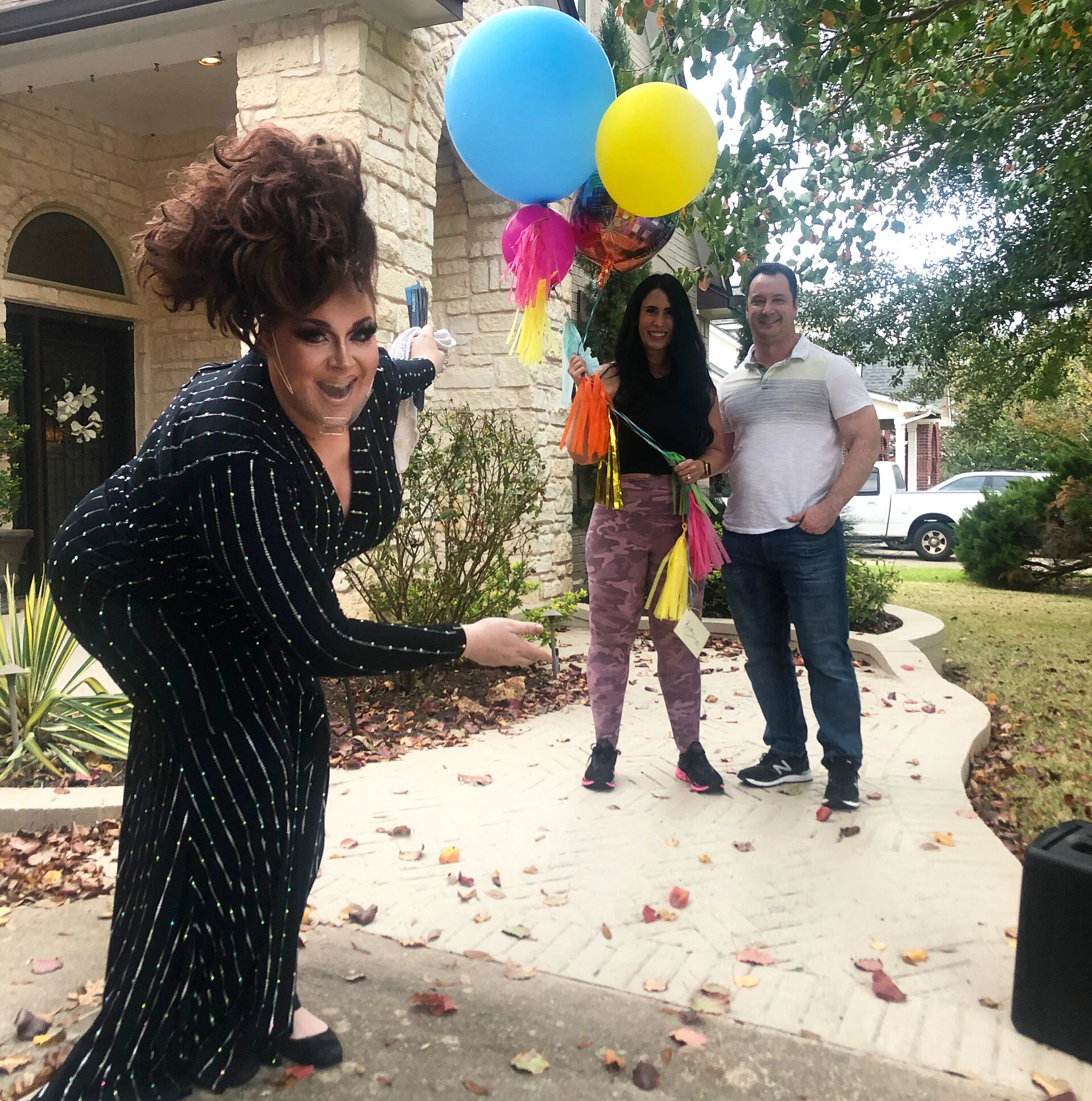Three people celebrating outdoors, with a woman holding colorful balloons, standing on a sidewalk leading to a house with a brick exterior and front yard with fallen leaves.