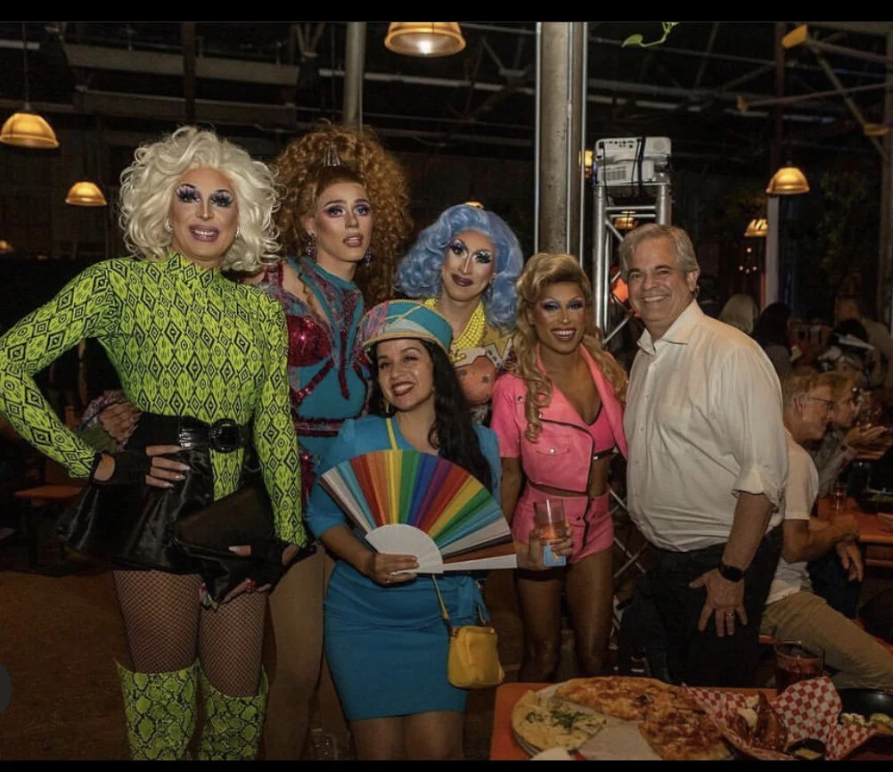 Group of six people, five drag performers in colorful costumes and a man in a white shirt, standing together at a social event with pizza on the table in front of them.