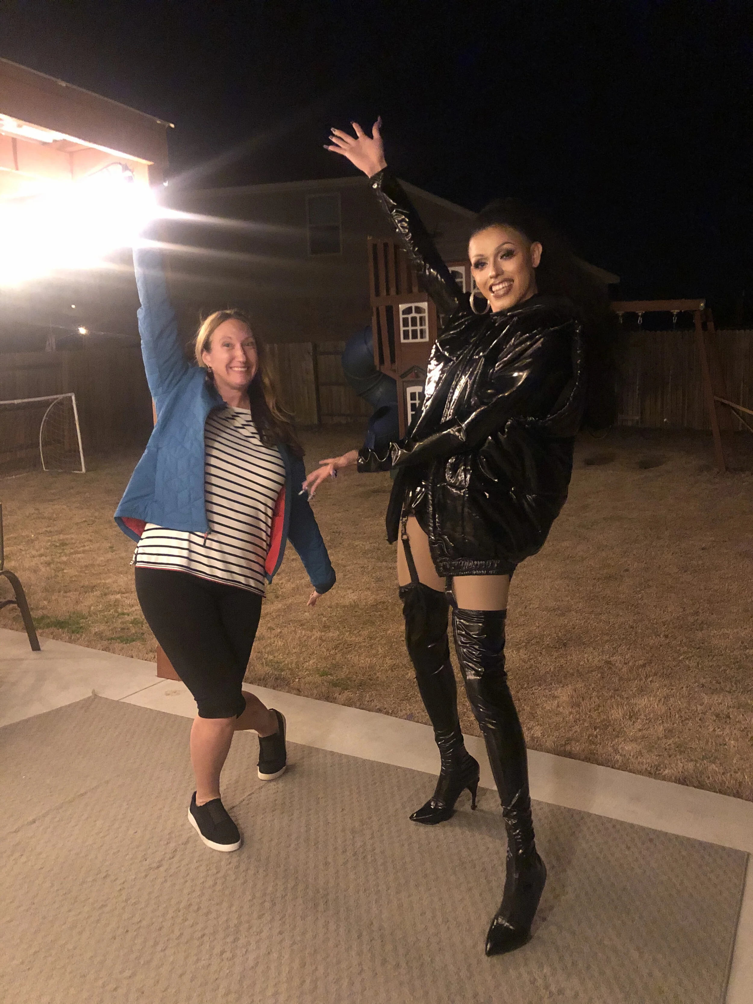 Two women, one in a blue jacket and striped shirt, the other in a shiny black leather outfit with thigh-high boots, are posing playfully outdoors at night in a backyard with play equipment in the background.