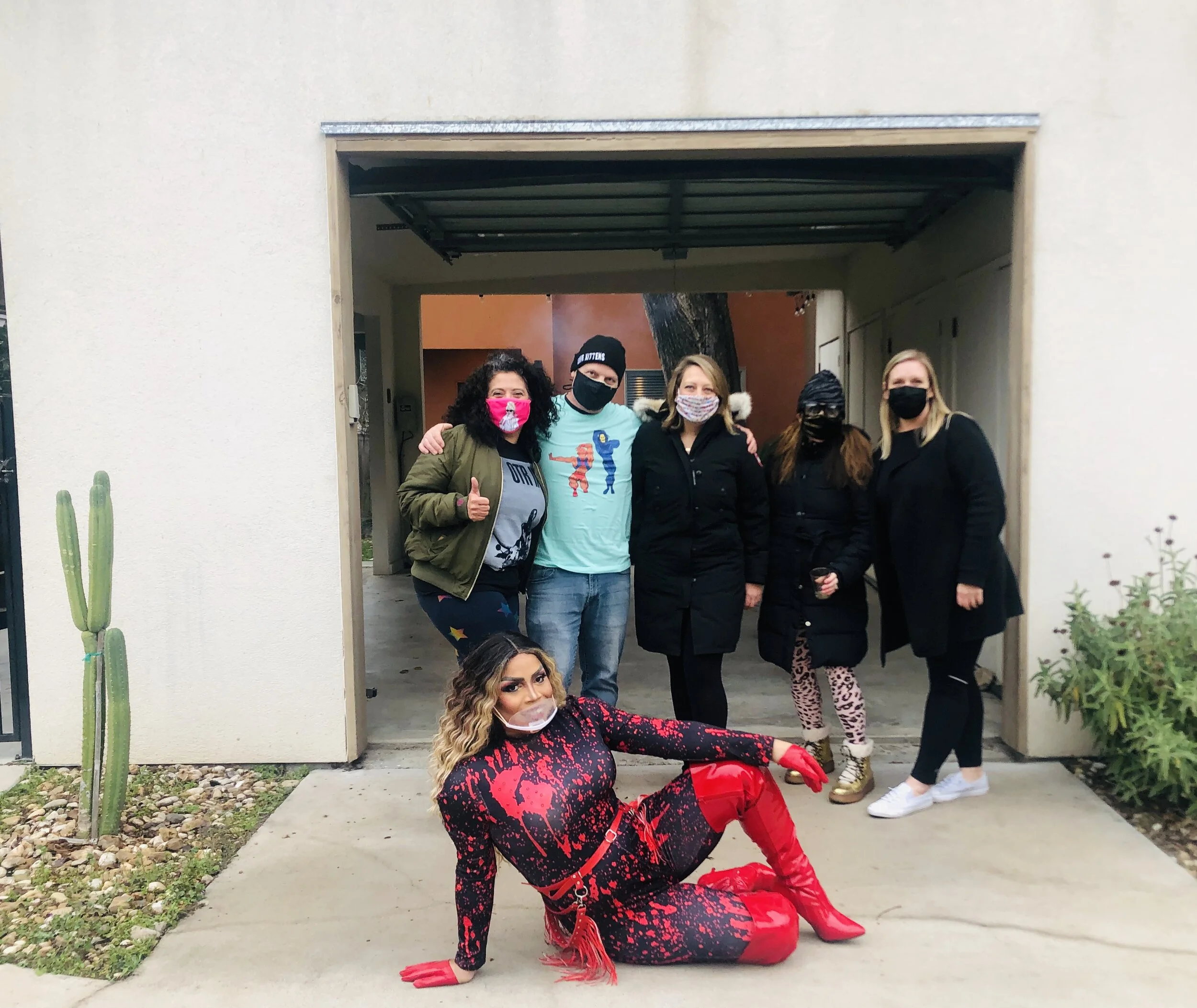 Group of six people wearing masks standing in a garage, with one person in a red and black outfit posing in front.